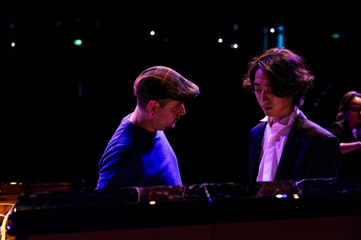 Two musicians collaborating at a piano on a dimly lit stage, one wearing a blue jacket and flat cap.