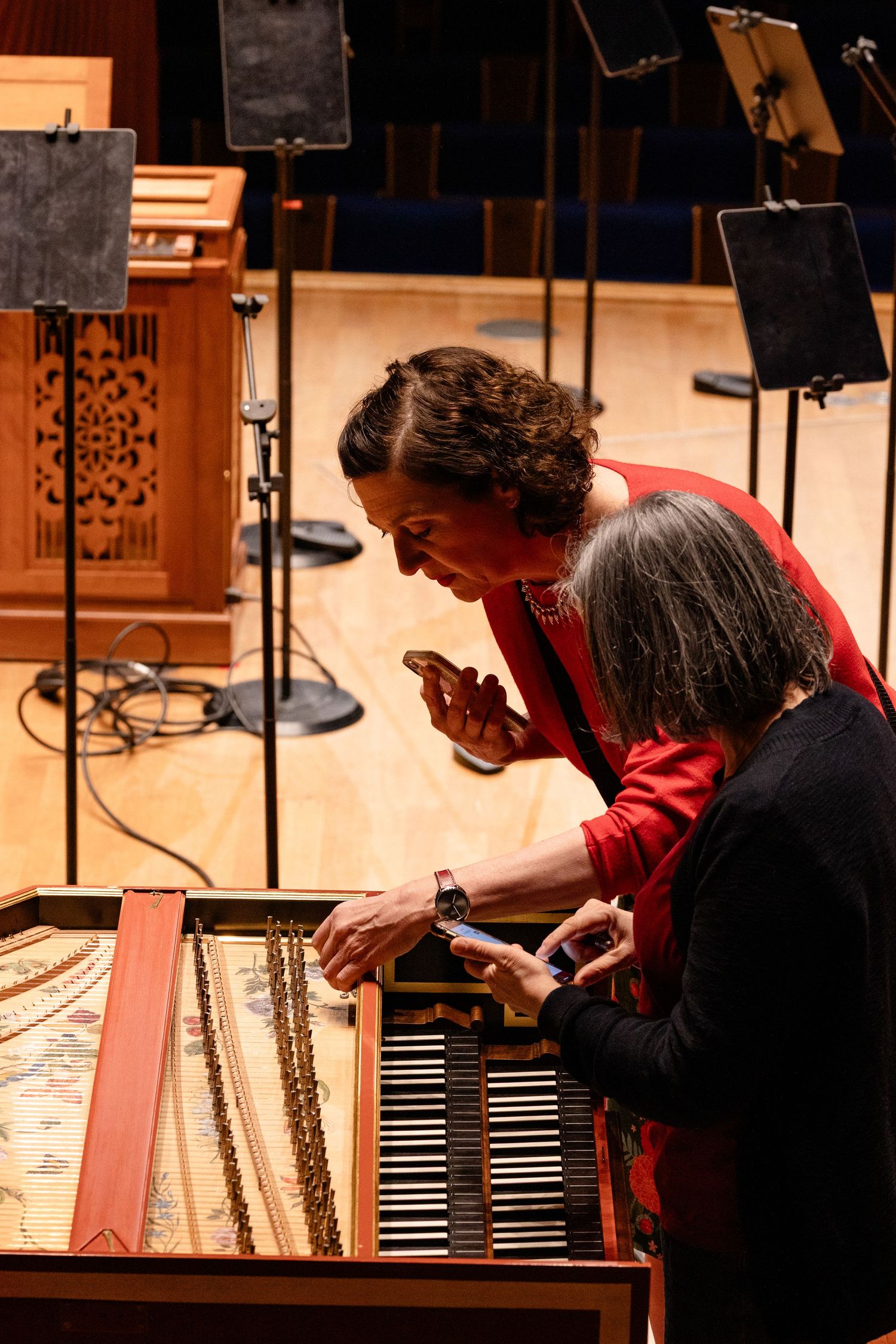 Two women examine the keys of a harpsichord on a concert hall stage, one holding a smartphone.