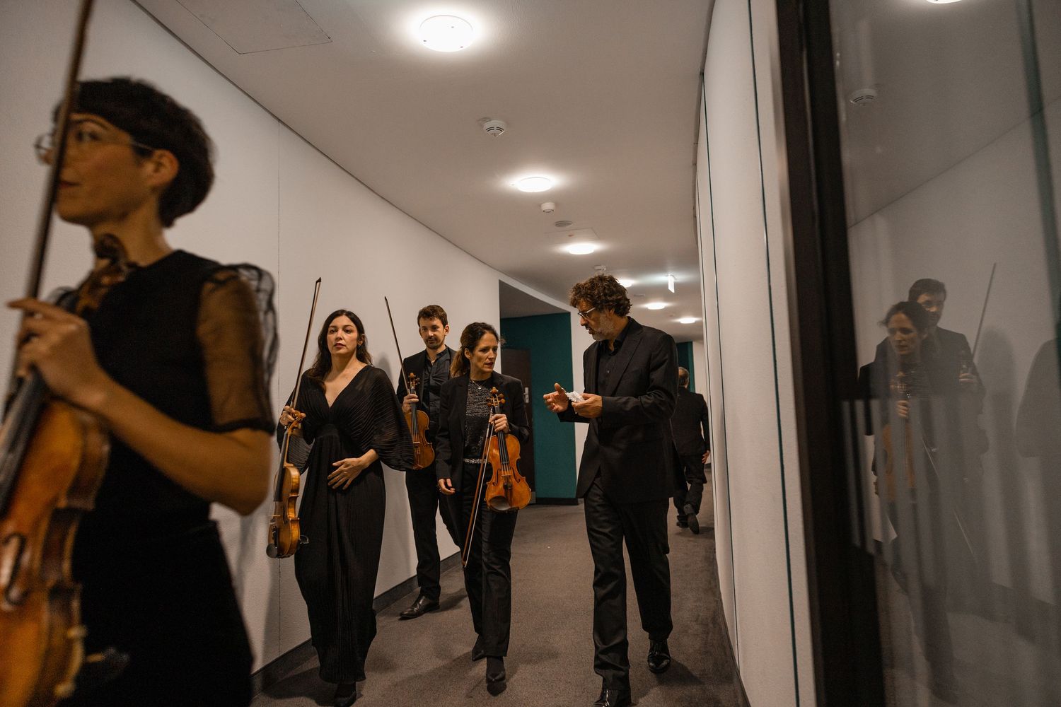 String quartet musicians in black attire carry violins through a modern corridor before a performance.