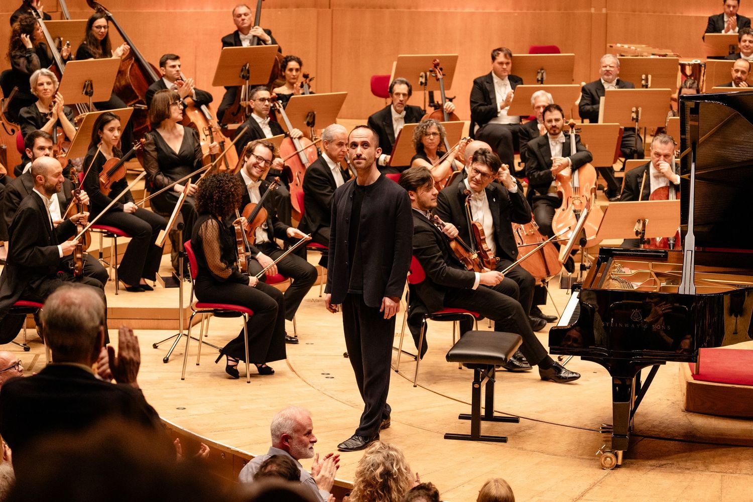 Orchestra performers take a bow on stage in a concert hall, with a grand piano visible and musicians holding instruments.