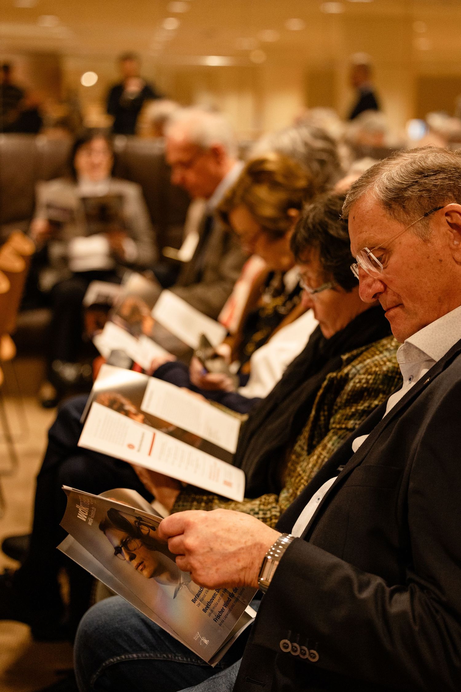 Audience members seated in rows reading printed programs at a formal conference or concert event.