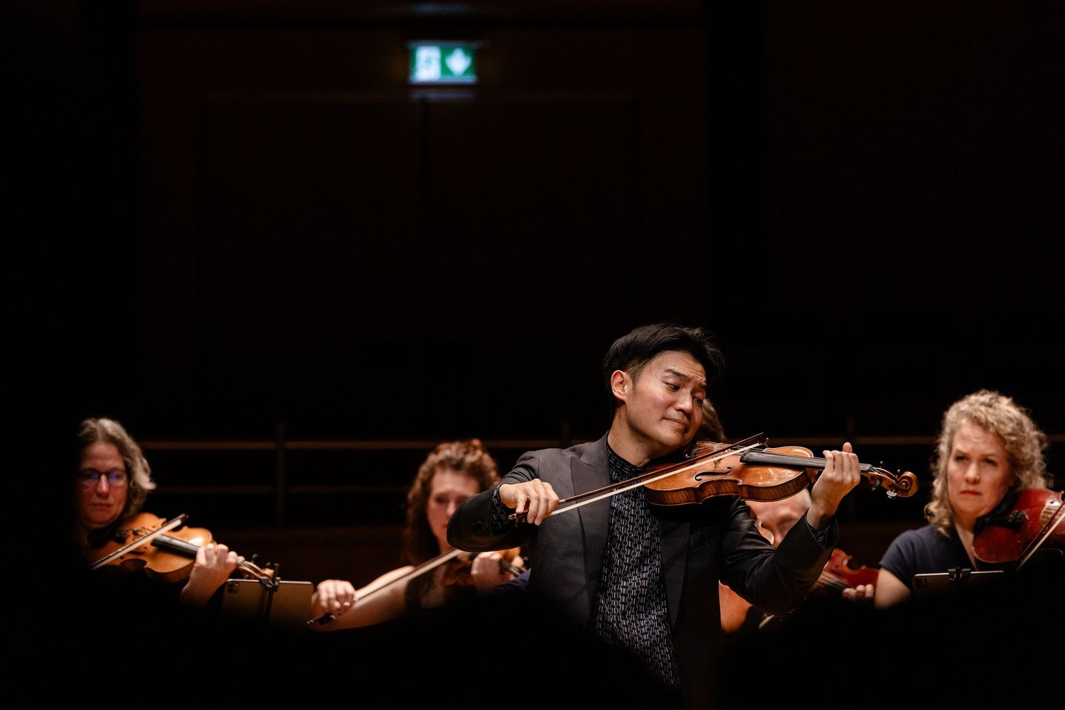 Young violinist performs passionately with orchestra musicians playing violins in a darkened concert hall.