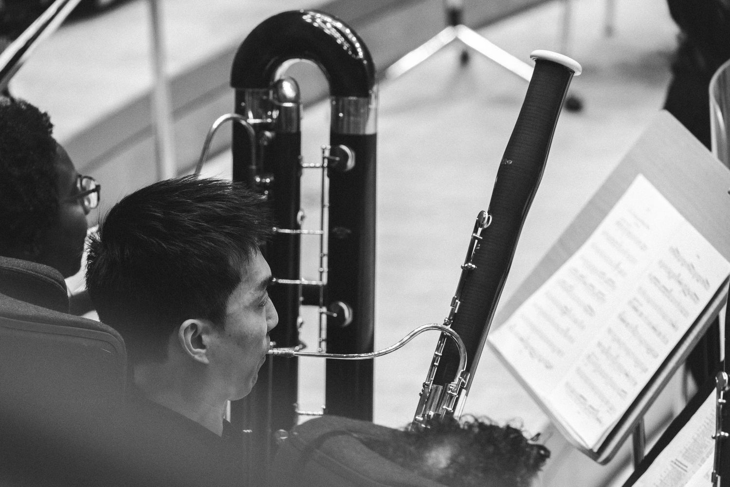 Black and white photo of a musician playing bassoon while reading sheet music in an orchestra rehearsal.
