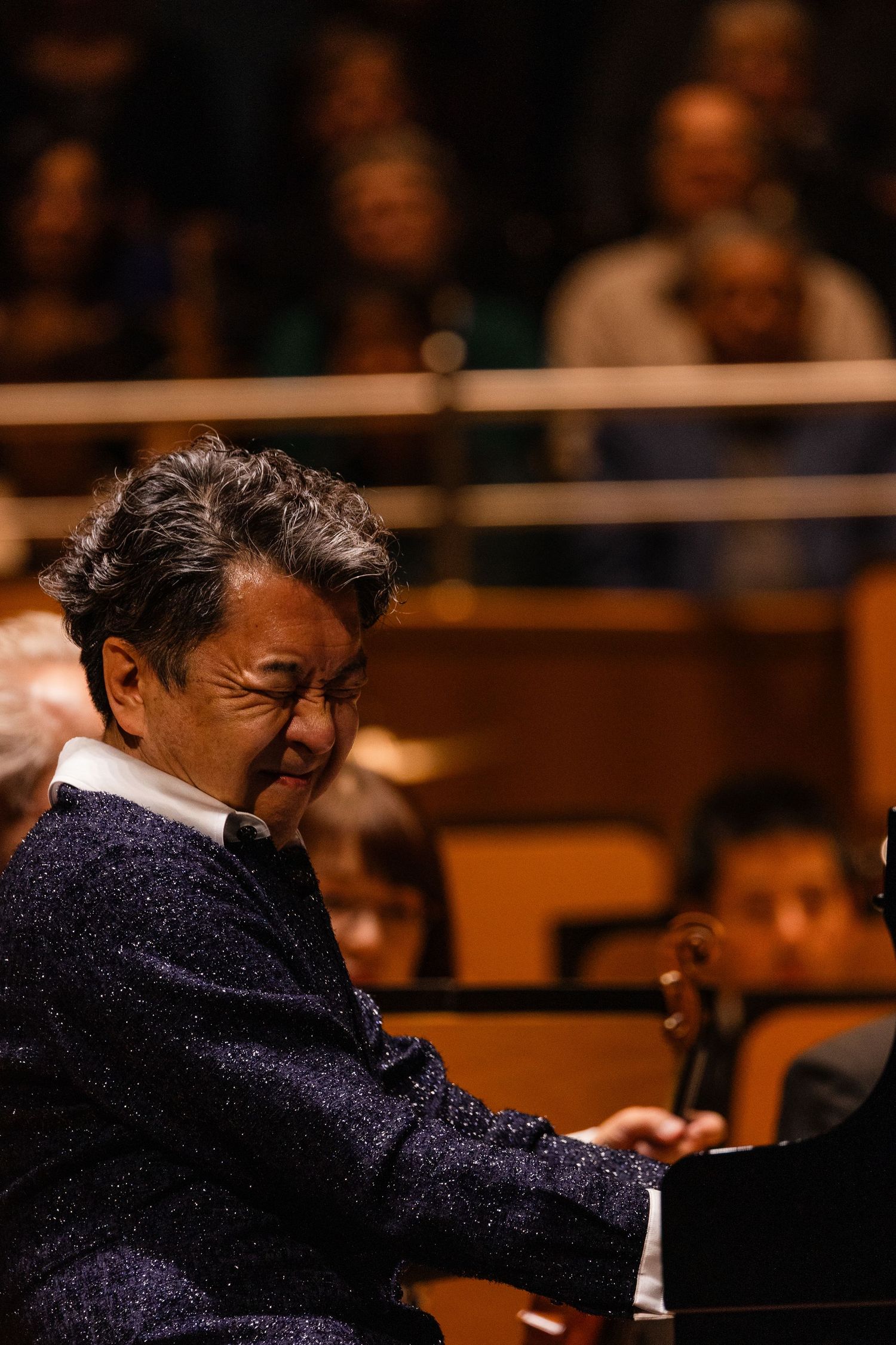 Pianist performing on stage in a sparkly blue jacket, smiling while playing, with an audience visible in the background.