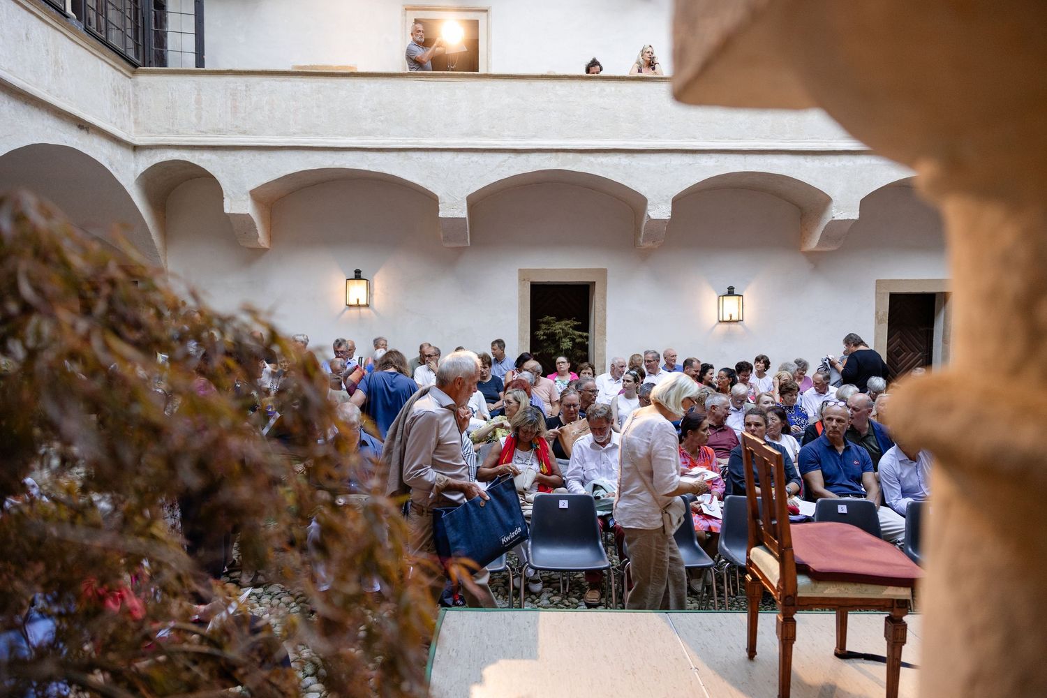 Large crowd gathered in a sunlit courtyard with arched architecture for an outdoor event or conference.