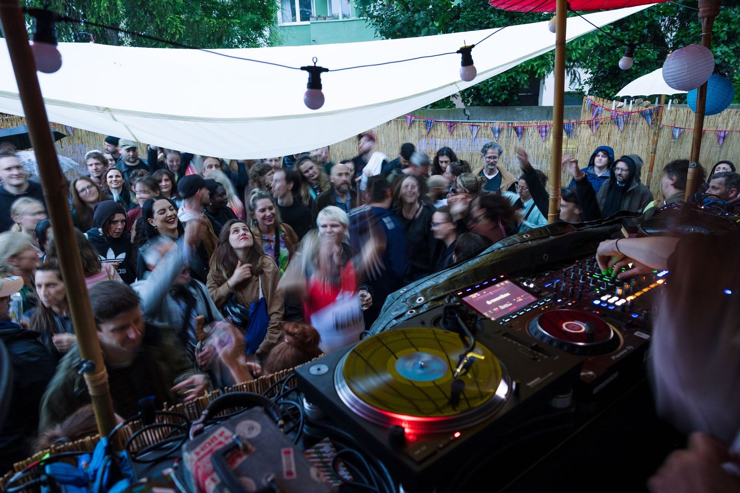 DJ performing at an outdoor party with a crowd of people dancing and celebrating under string lights.