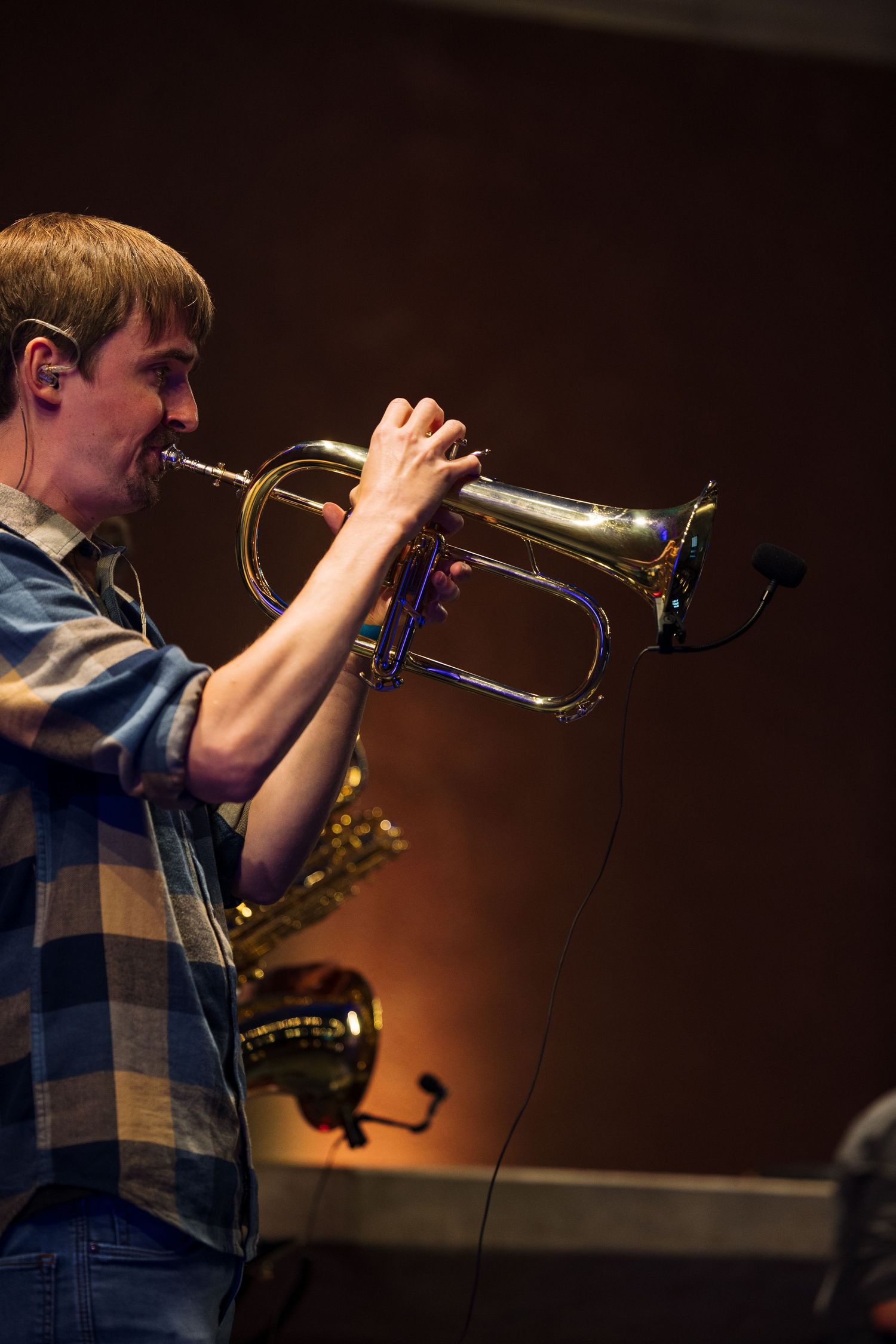 Young musician playing a brass trumpet on stage under warm lighting, wearing a plaid shirt.