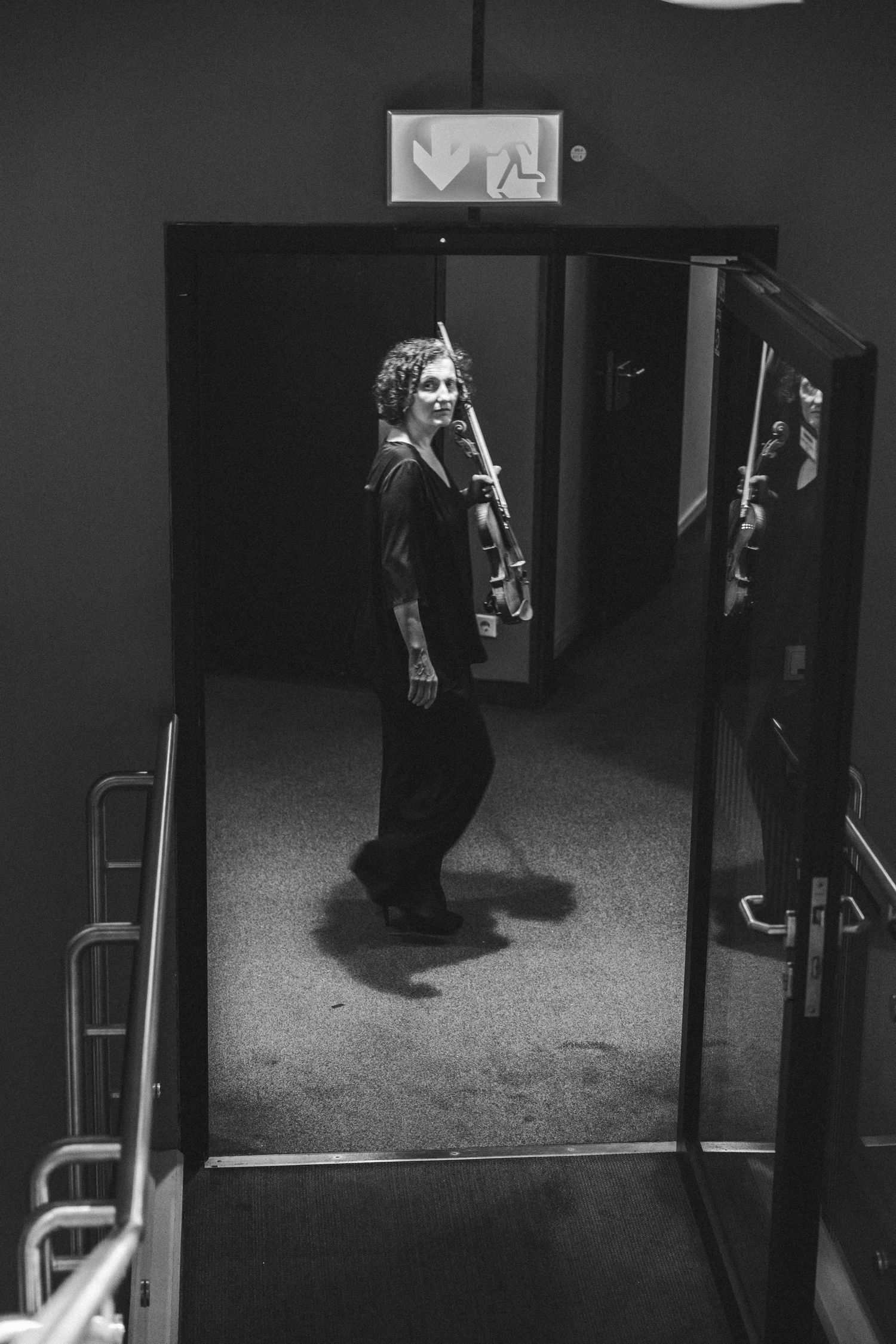 Black and white photo of a woman holding a violin walking through a doorway under an exit sign.