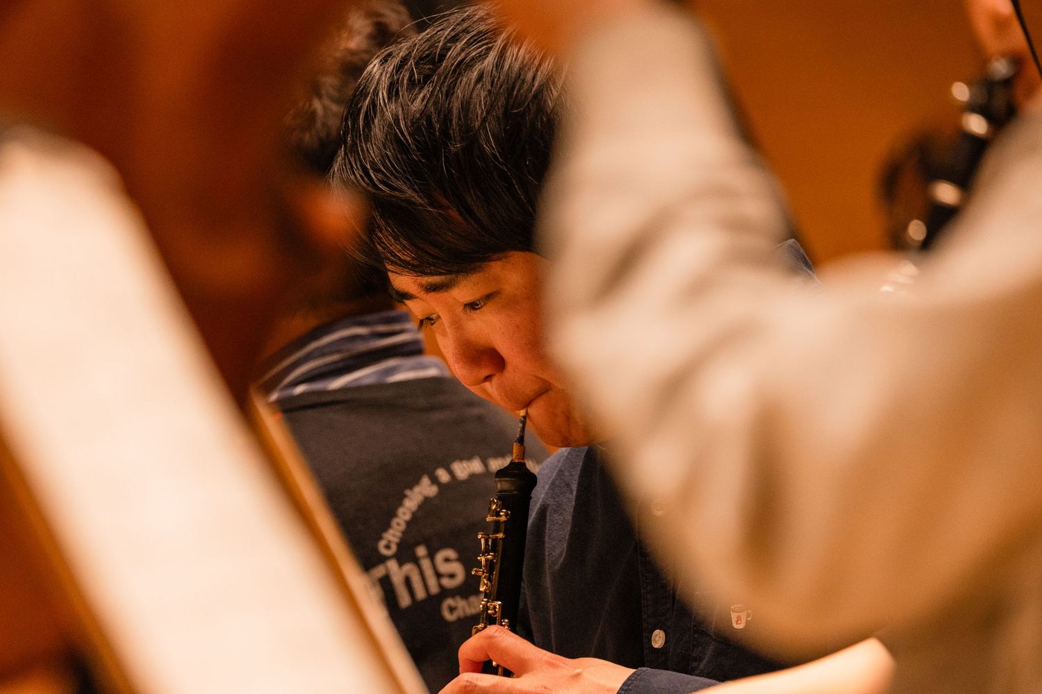 Musician playing clarinet during orchestra rehearsal, focused on sheet music in warm concert hall lighting.