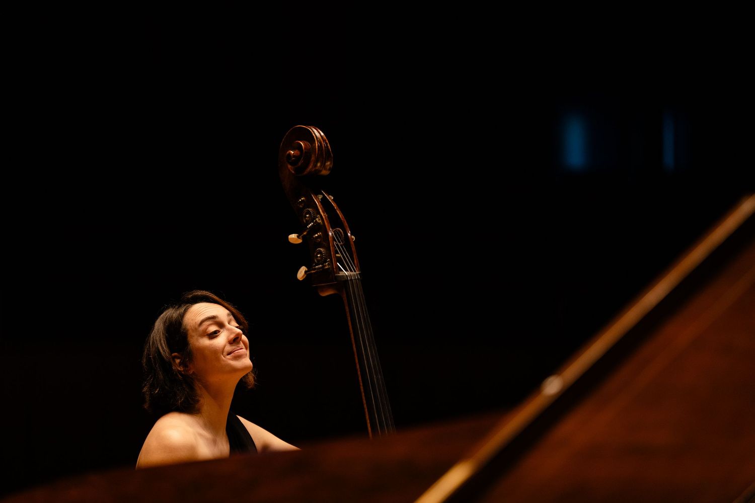A woman plays cello on a dimly lit concert stage, her face illuminated against a dark background.