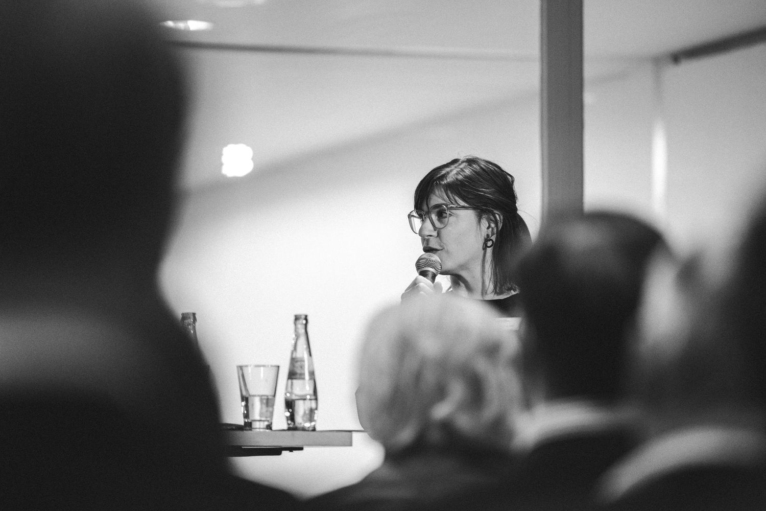 Black and white photo of a woman with glasses listening attentively during an indoor conference or meeting.