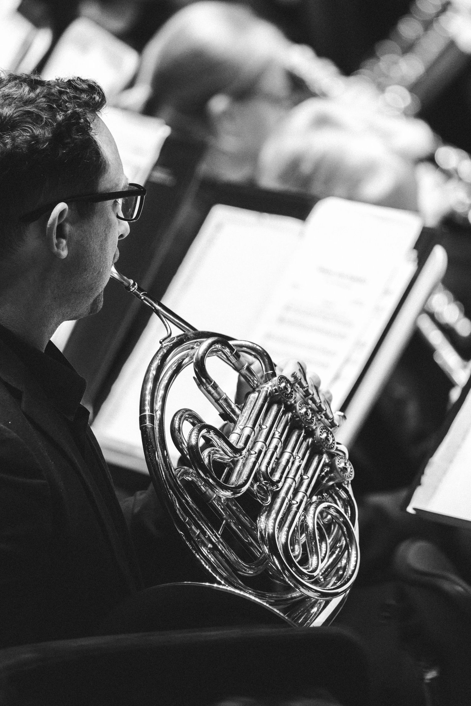 Black and white photo of a musician playing a French horn while reading sheet music in an orchestra setting.