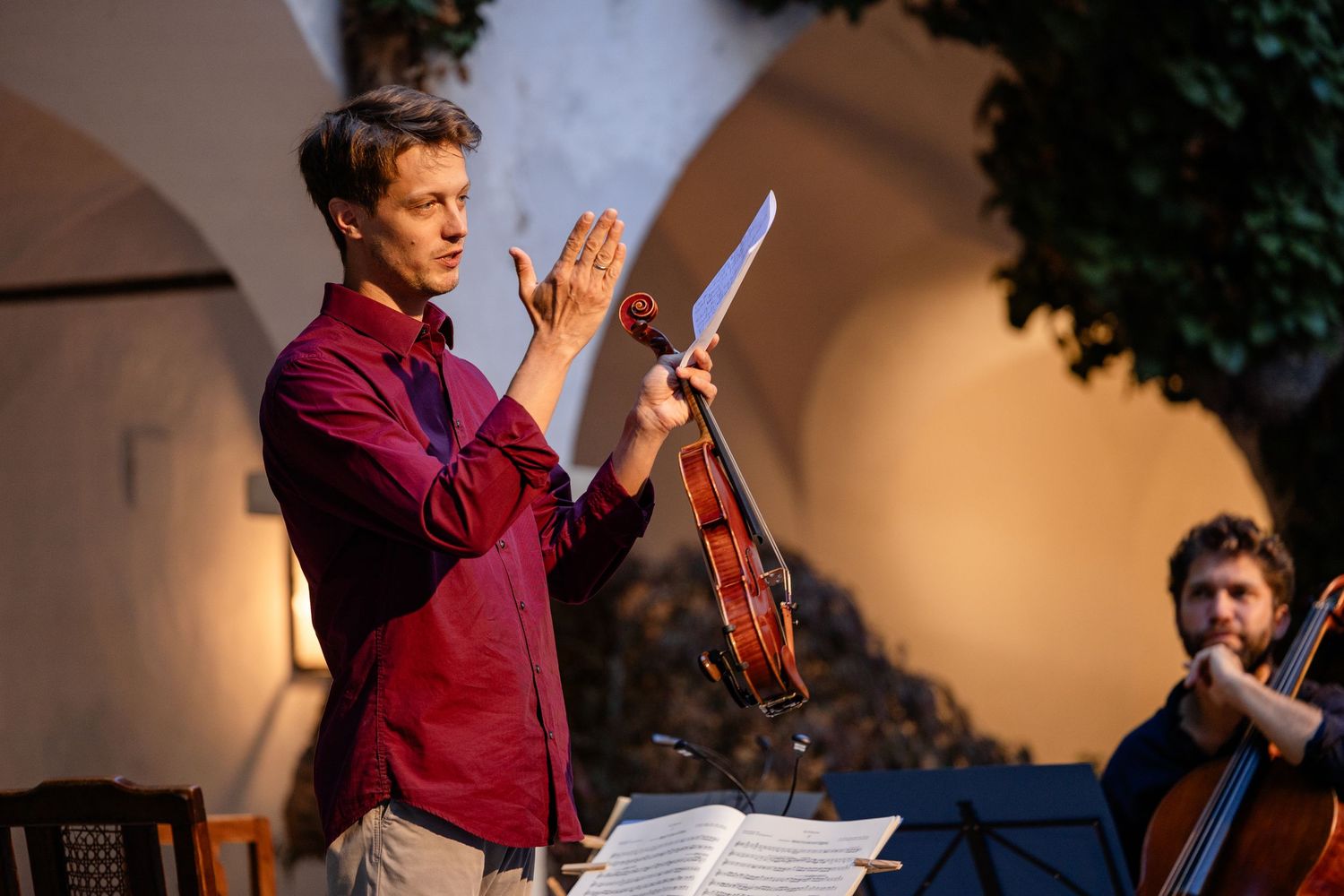 Conductor holding violin and sheet music gestures during outdoor classical music performance with cellist in background.