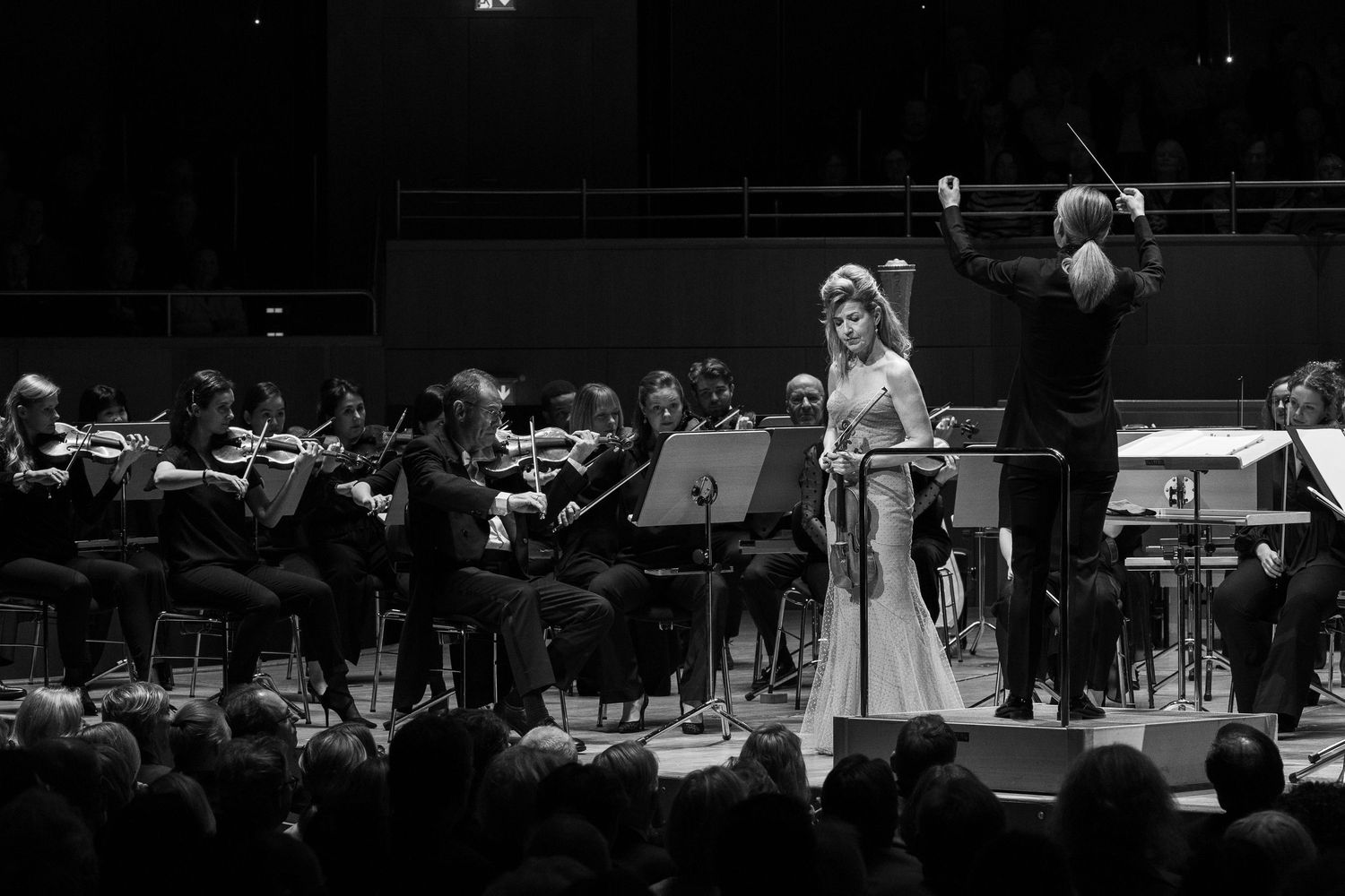 Black and white photo of a female violinist performing with a full orchestra, conductor raising baton on stage.