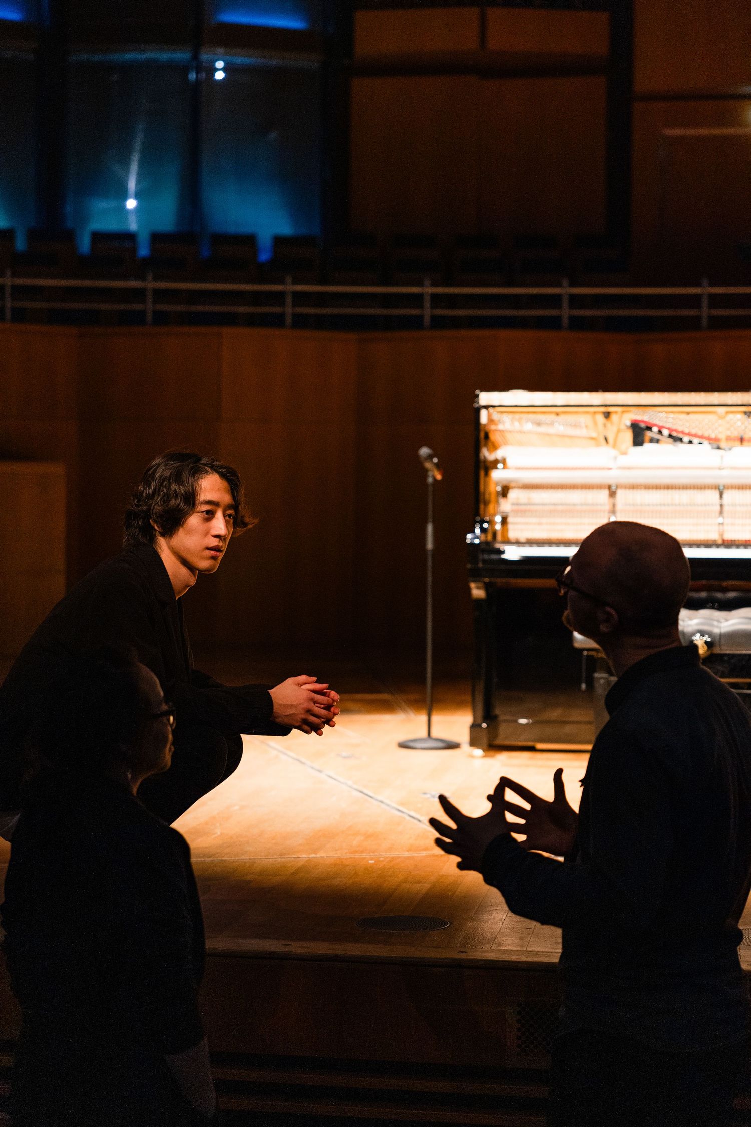 Two musicians converse on a concert hall stage near a grand piano under warm dramatic lighting.