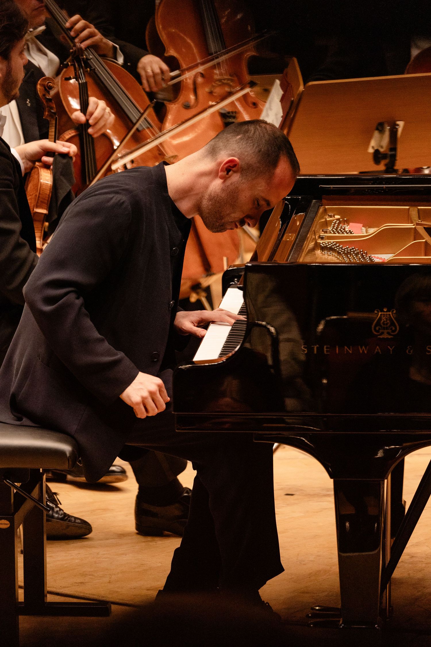 A male pianist performs intensely on a Steinway grand piano with an orchestra behind him in a concert hall.
