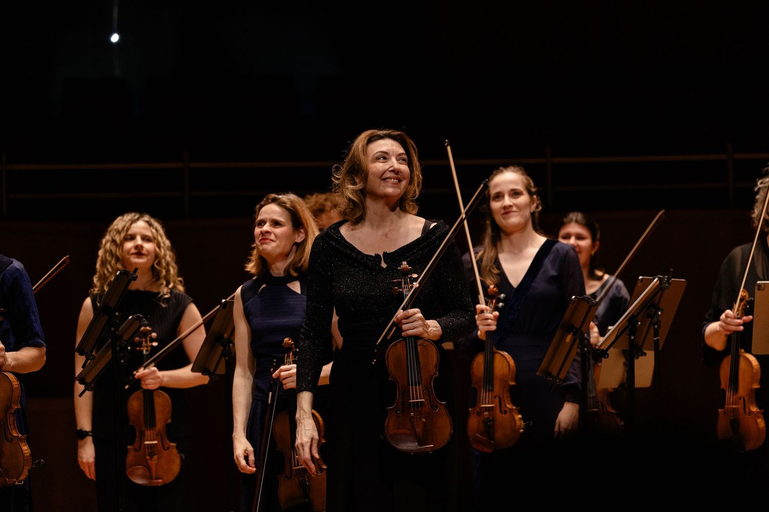Female violinists in navy dresses hold instruments on stage, with a central performer holding a bow at a classical music concert.