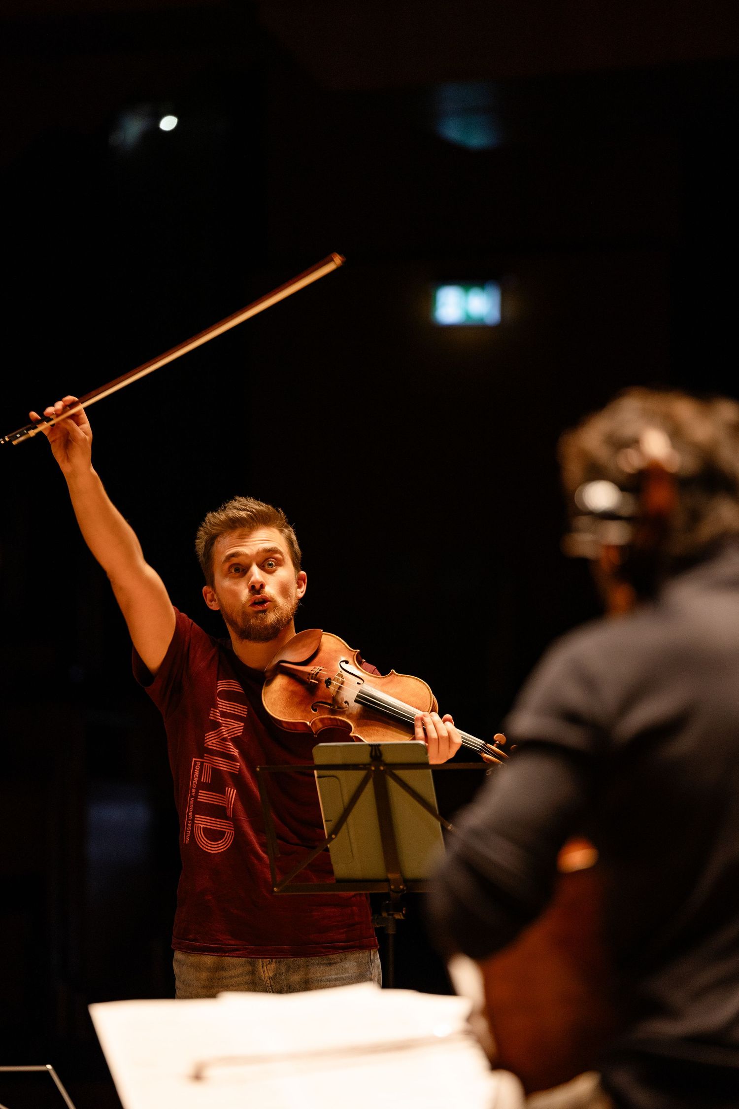 Violinist in red shirt raises bow dramatically while reading sheet music on stage during rehearsal.