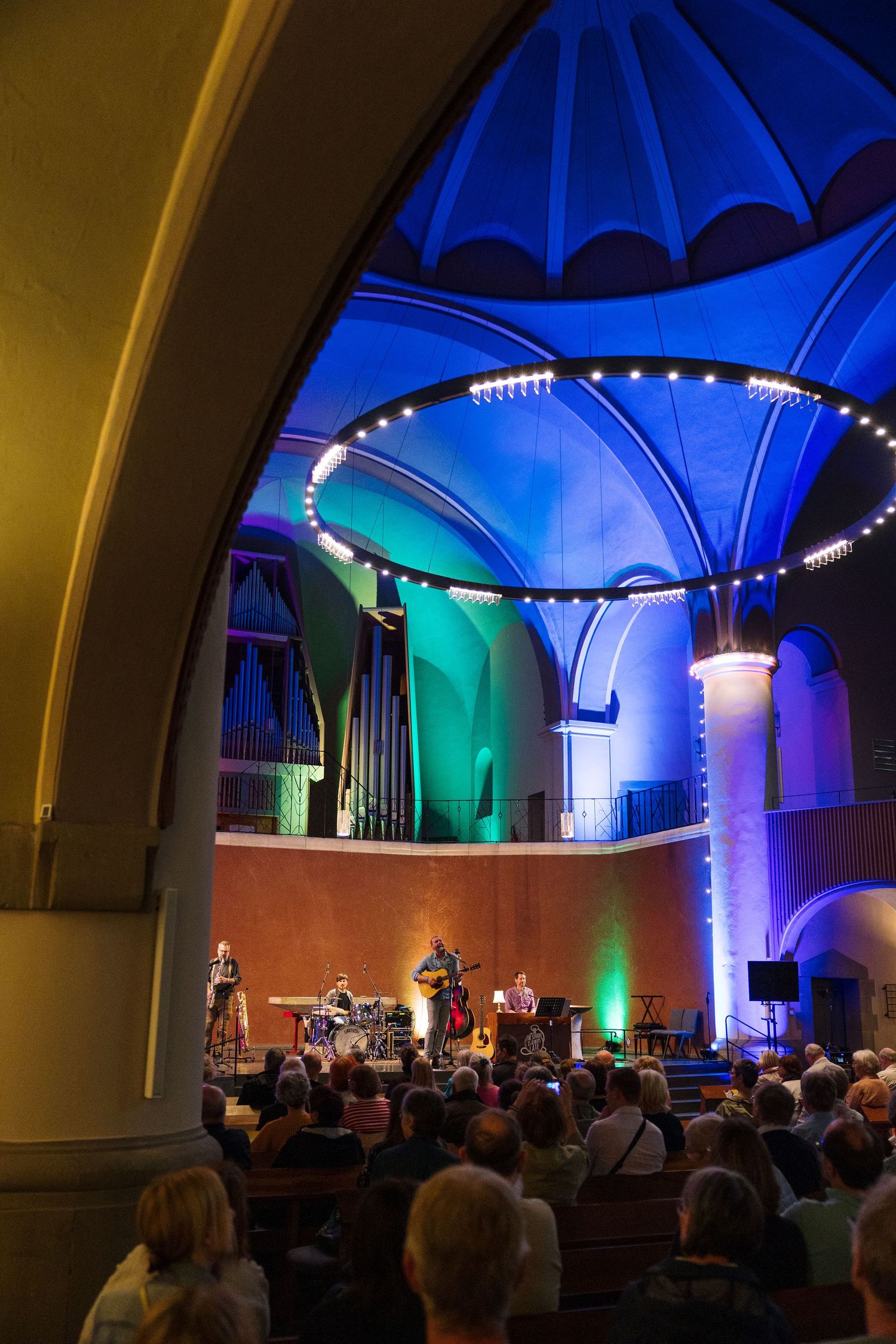 Live concert in a stunning church venue with colorful blue and green lighting illuminating vaulted ceilings above an audience.