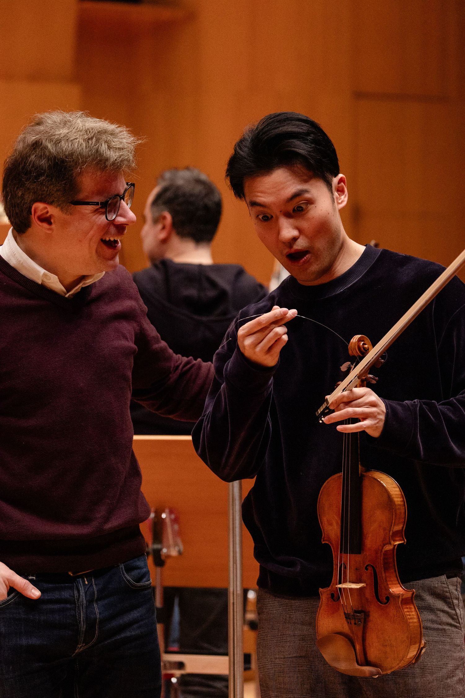 Two musicians discuss violin technique in a concert hall, one holding a violin and bow during rehearsal.