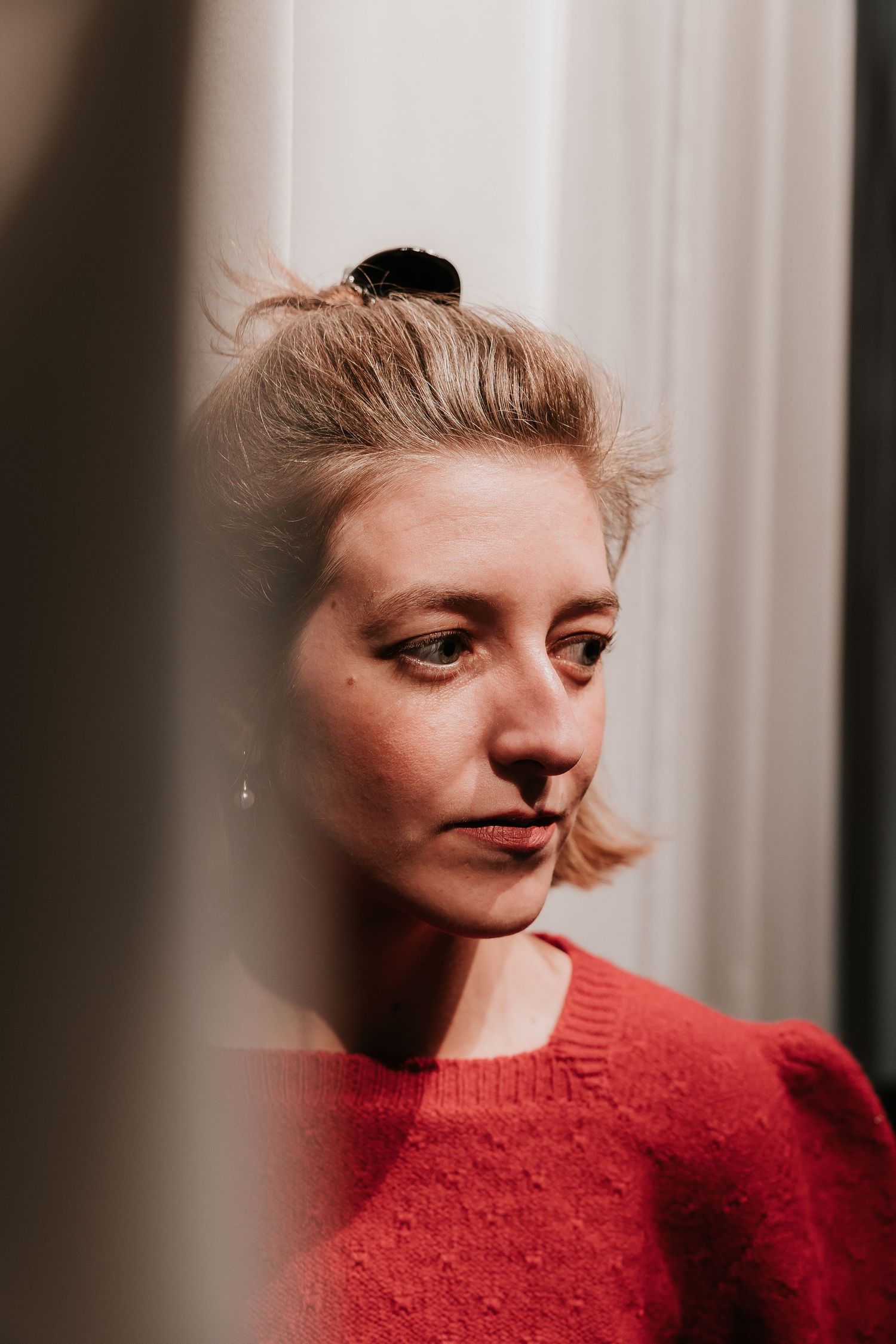 A pensive young woman with blonde hair in a bun wearing a red sweater gazes downward in soft natural light.