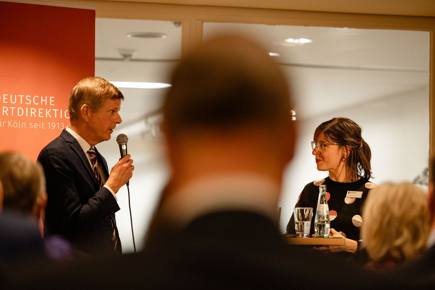 Man speaking into microphone at Deutsche Sportdirektion event, engaging with a woman in a professional indoor setting.
