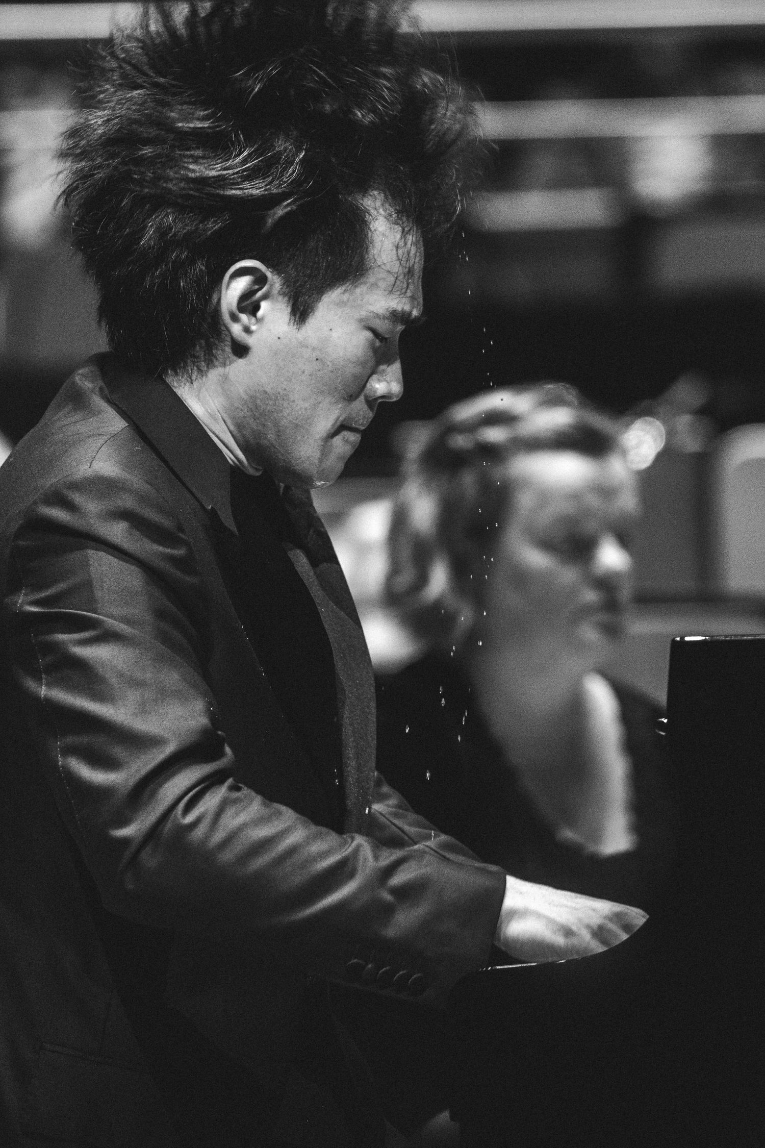 Black and white photo of a male pianist performing passionately at a grand piano, hair disheveled mid-play.