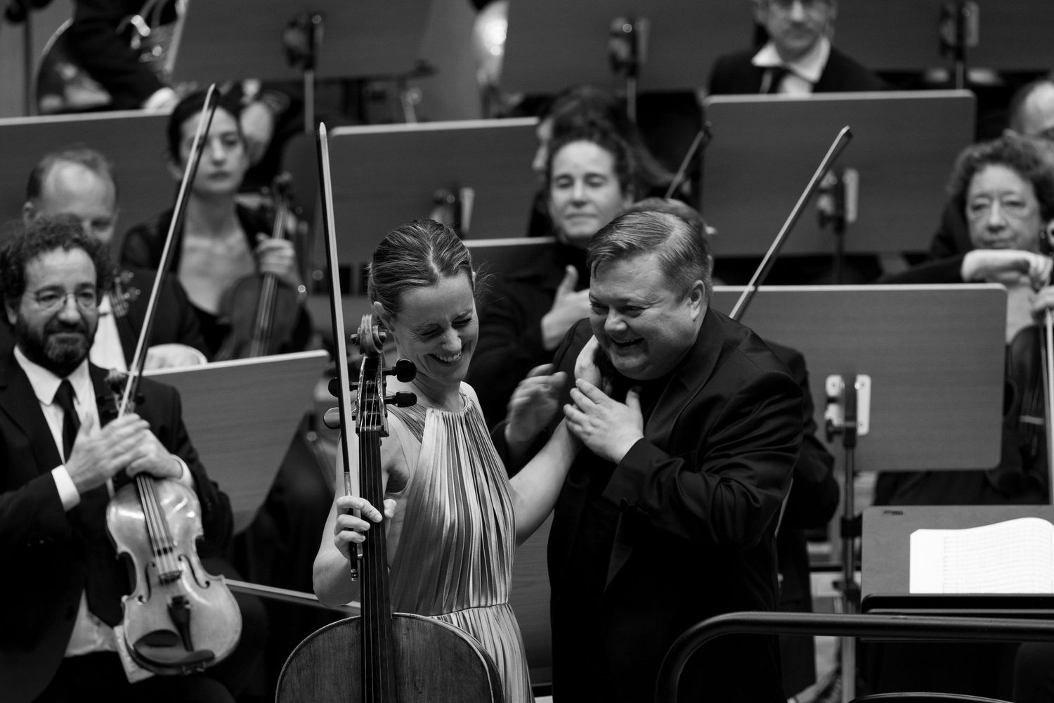 Black and white photo of musicians on stage, cellist and conductor sharing a moment during orchestral performance.