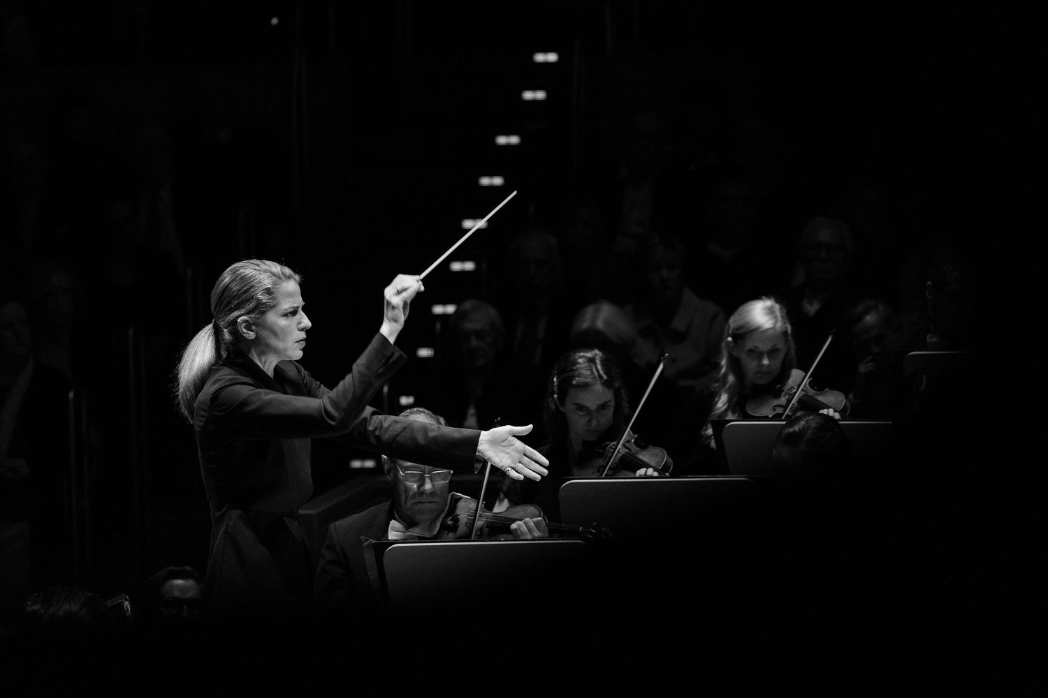 Black and white photo of a female orchestra conductor raising her baton while musicians play in the background.