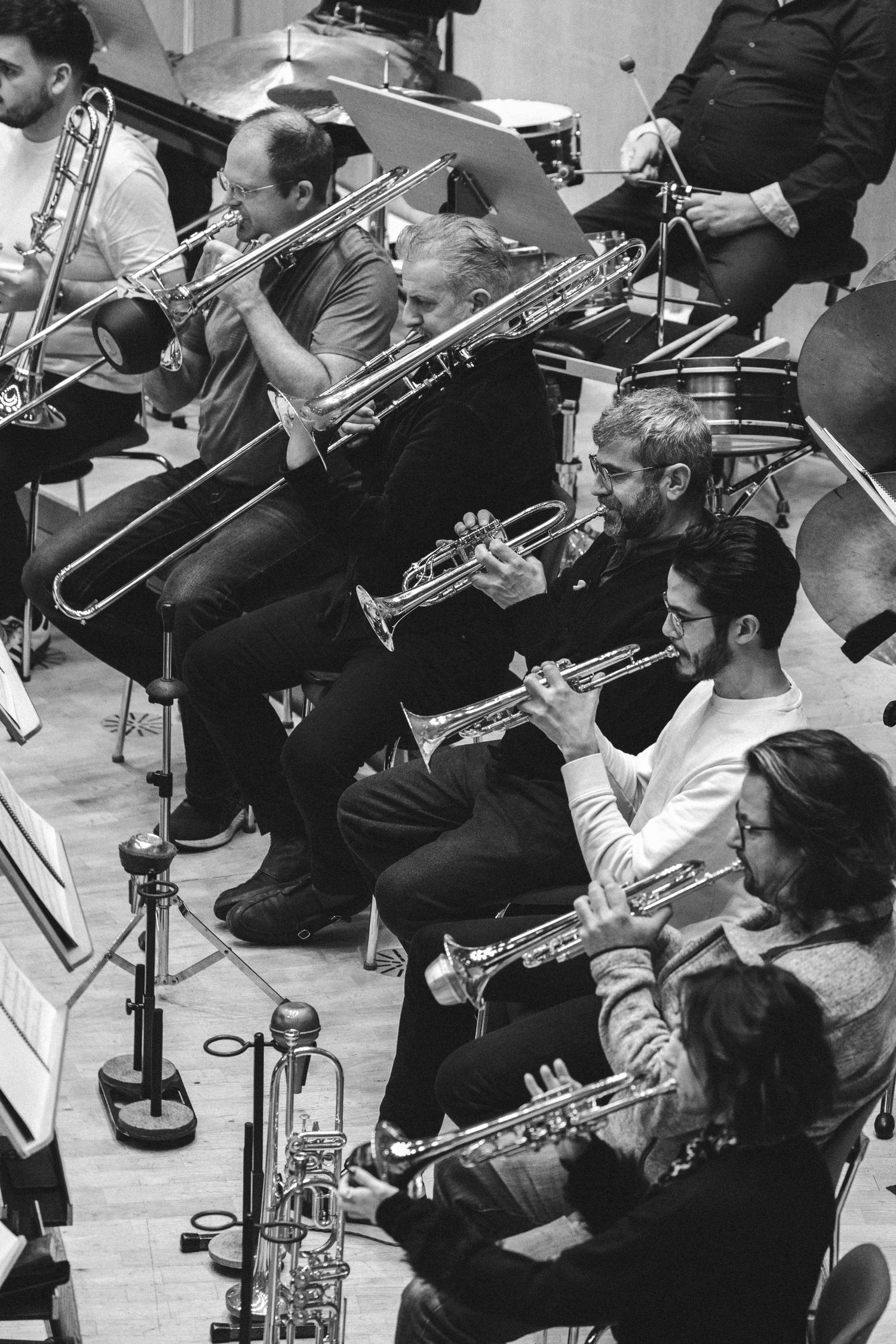 Black and white photo of orchestra musicians playing brass instruments including trumpets and trombones during rehearsal.