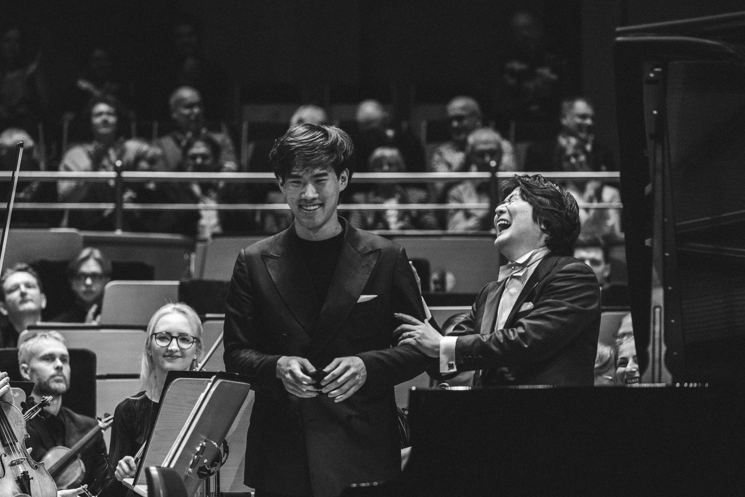 Two male musicians bow on stage with an orchestra in a black and white concert hall performance photo.