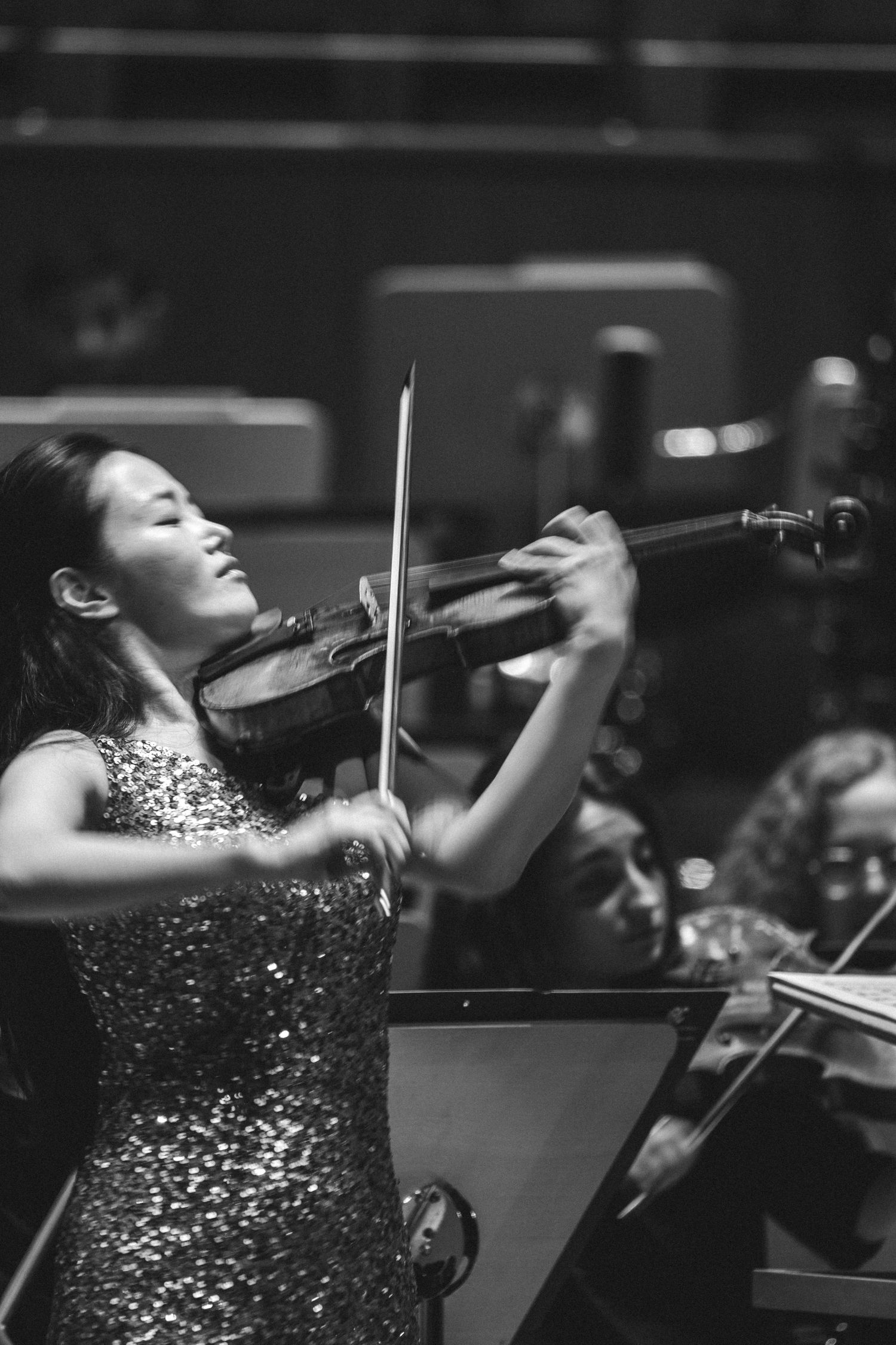A woman in a sequined dress plays violin with eyes closed in a black and white orchestra performance photo.