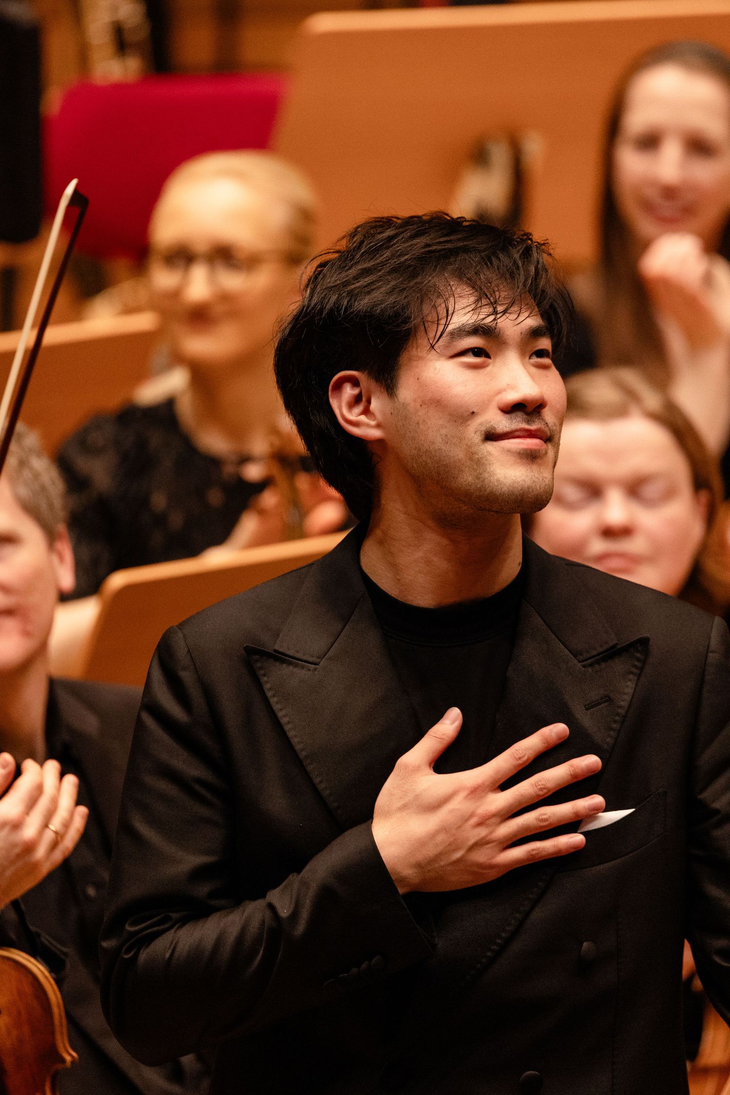 Young male musician in black jacket places hand on chest, bowing gracefully before an applauding orchestra.