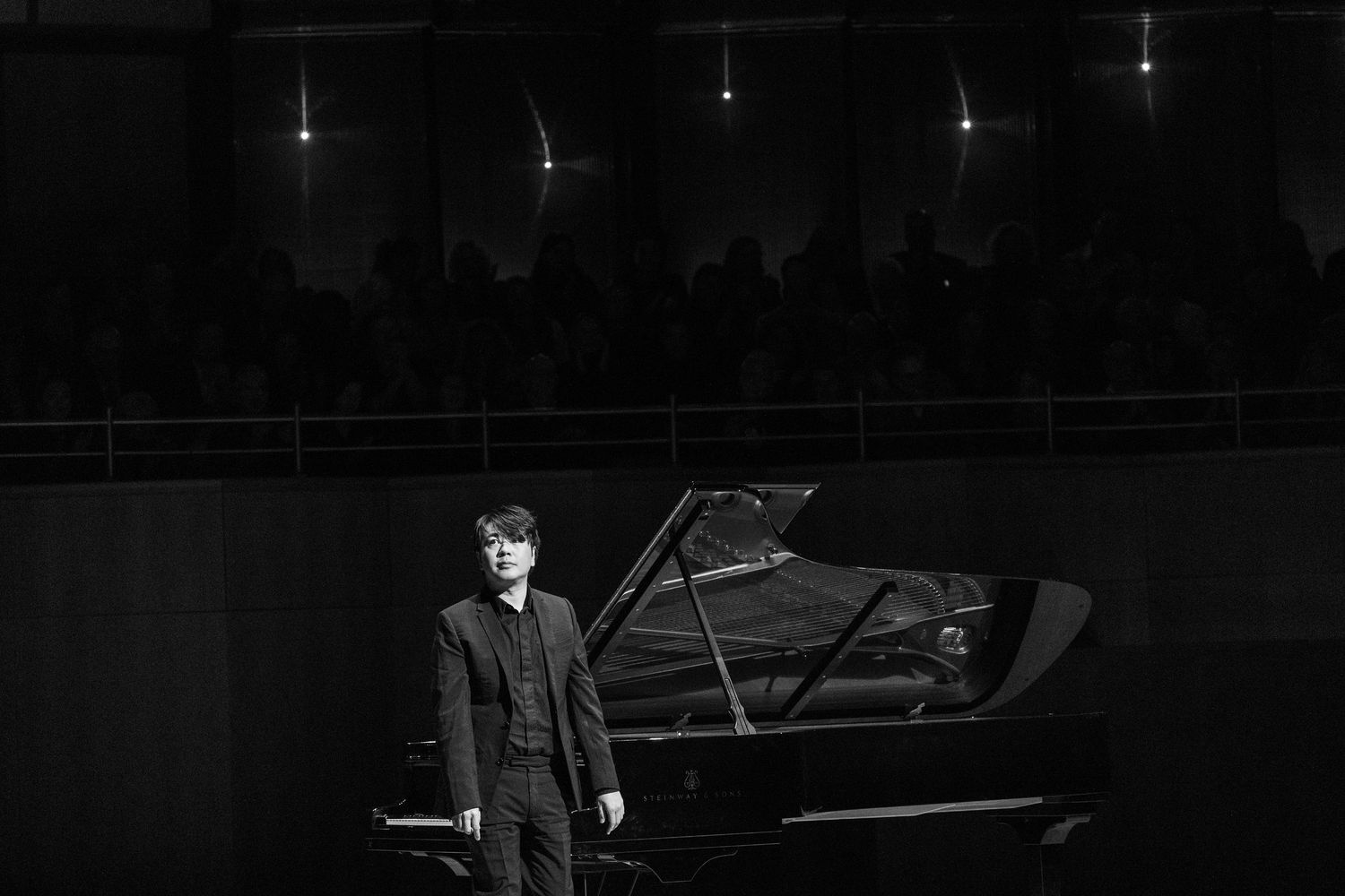 Black and white photo of a pianist standing beside a grand piano on a concert stage with audience in background.