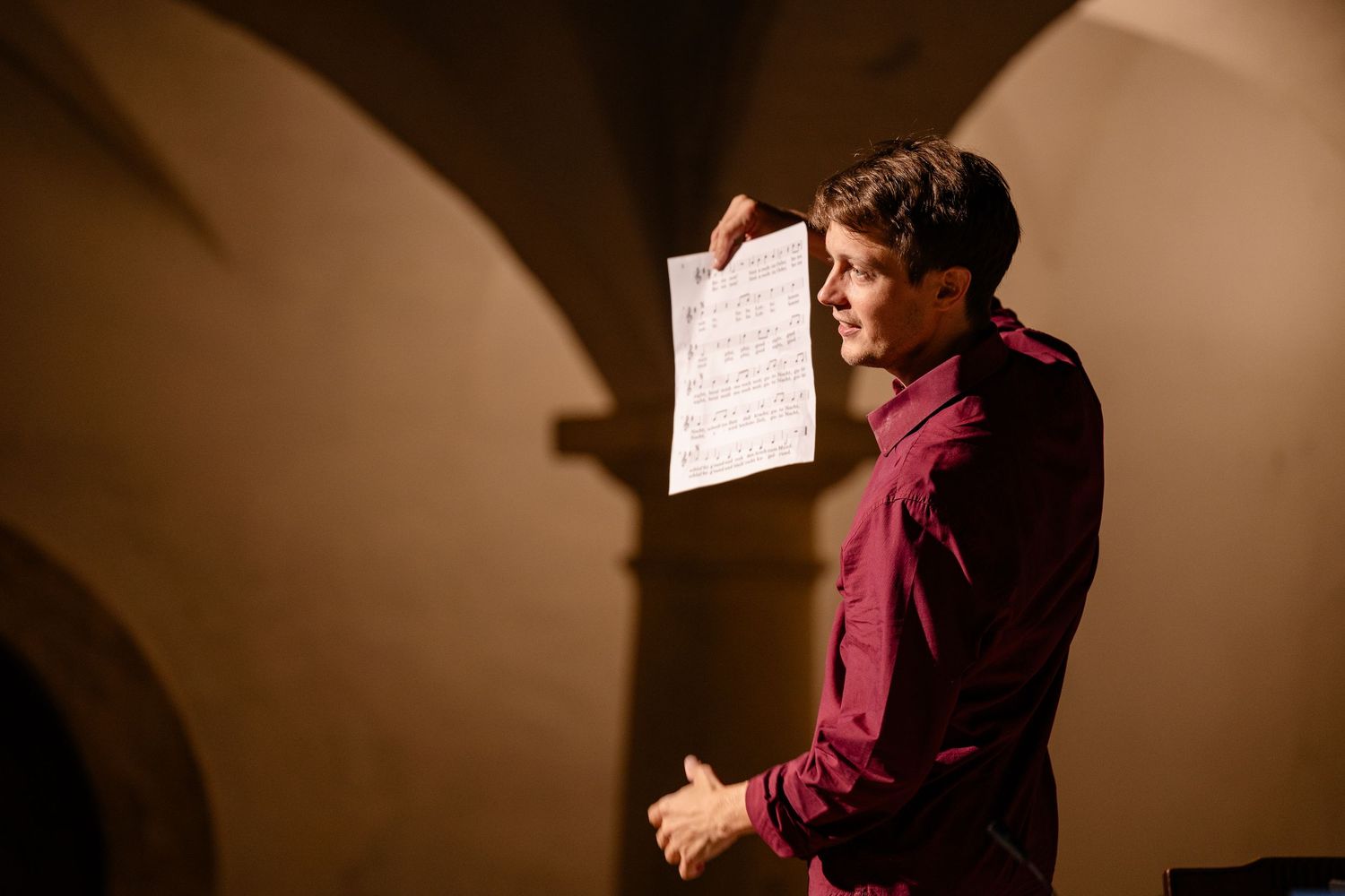 Young man in burgundy shirt holding sheet music under arched stone ceiling in dimly lit venue.