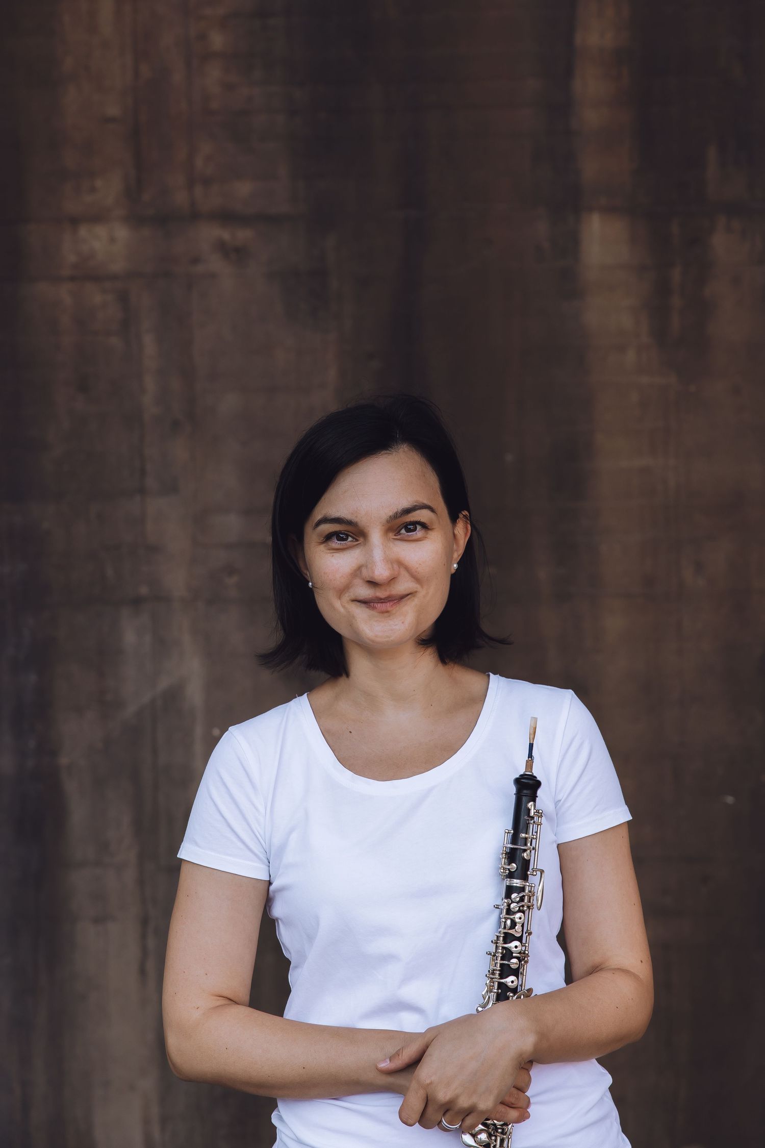 Woman in white t-shirt smiling while holding an oboe against a dark brown textured wall background.