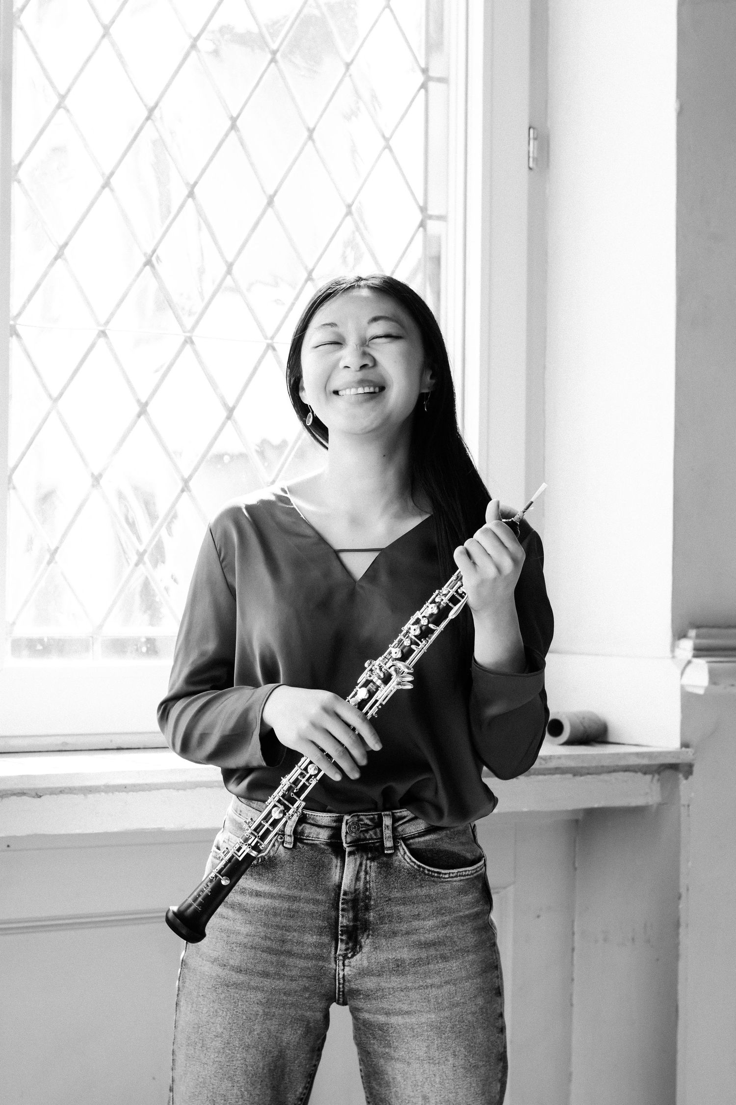 Young woman smiling and holding an oboe near a window in a black and white portrait photo.
