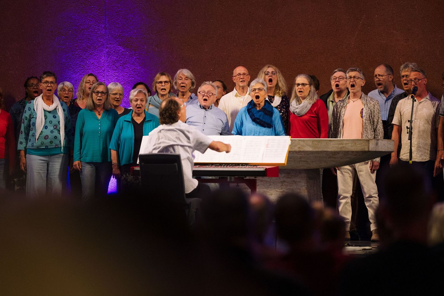 A large choir performs on stage with a conductor at a keyboard during a live choral concert event.