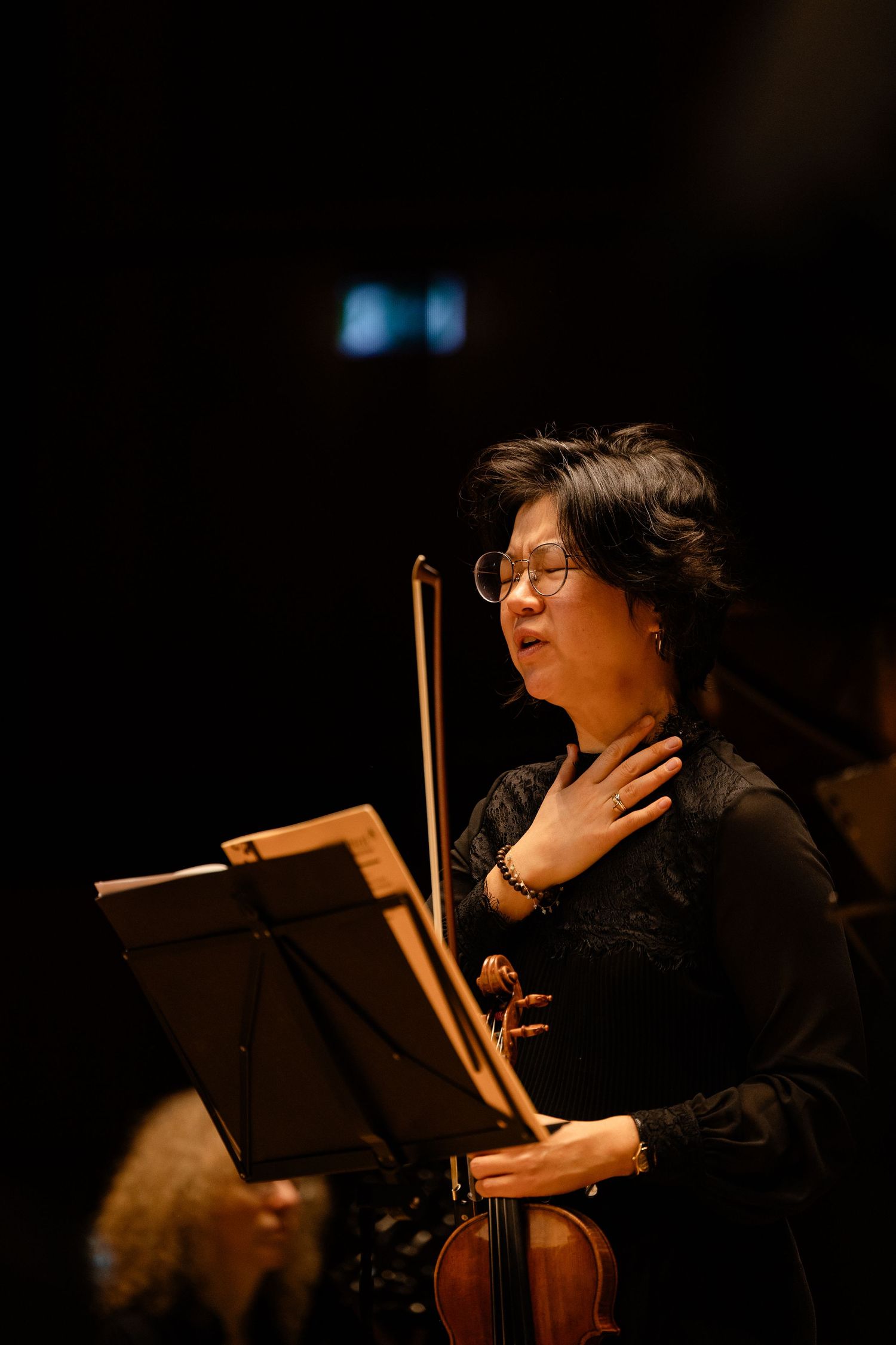 Female violinist in black attire holds violin and bow, standing at music stand during orchestral performance.