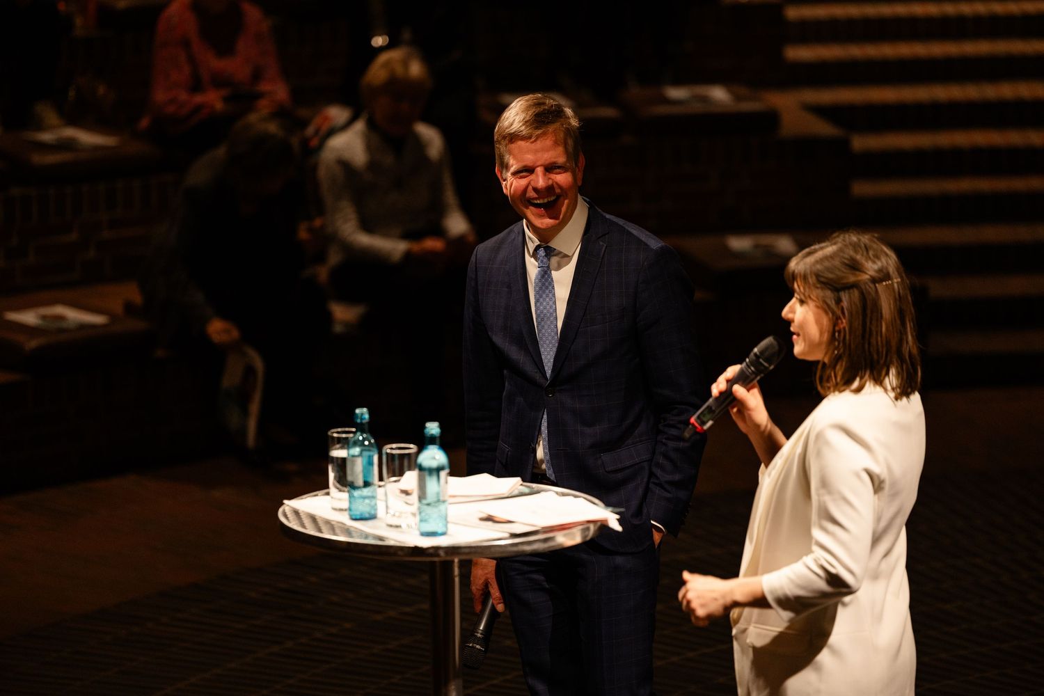 Man in blue suit and woman in white jacket speak at podium with microphone on stage at formal event.