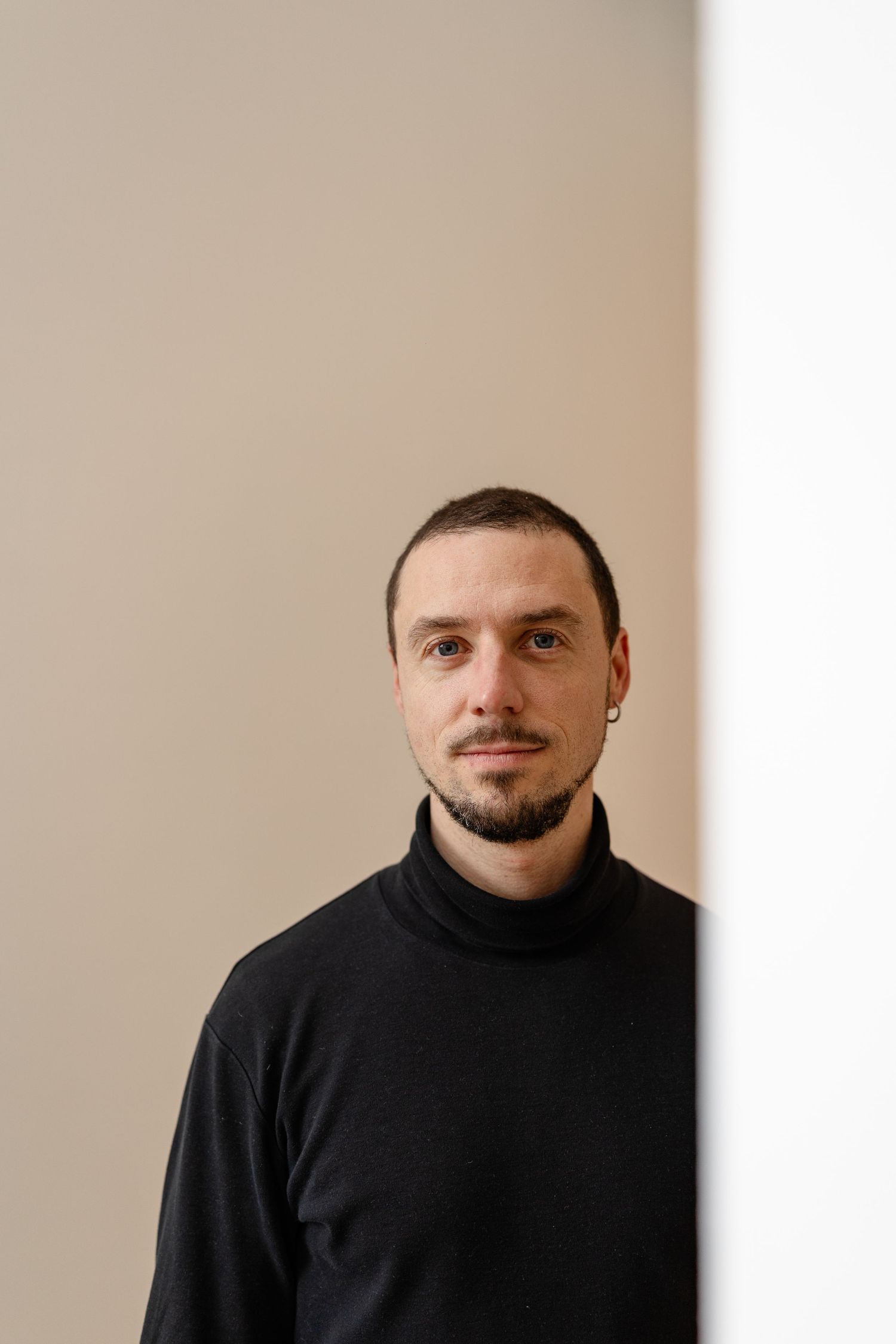 A young man with a goatee wearing a black turtleneck sweater stands against a beige wall.