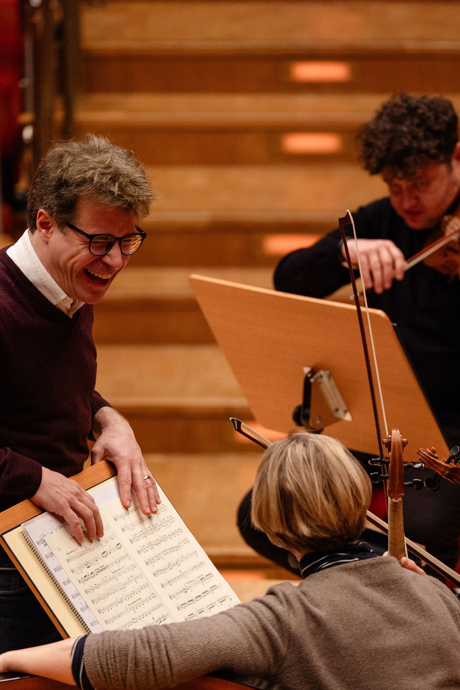 Smiling musician reviews sheet music with colleagues during an orchestra rehearsal in a concert hall.