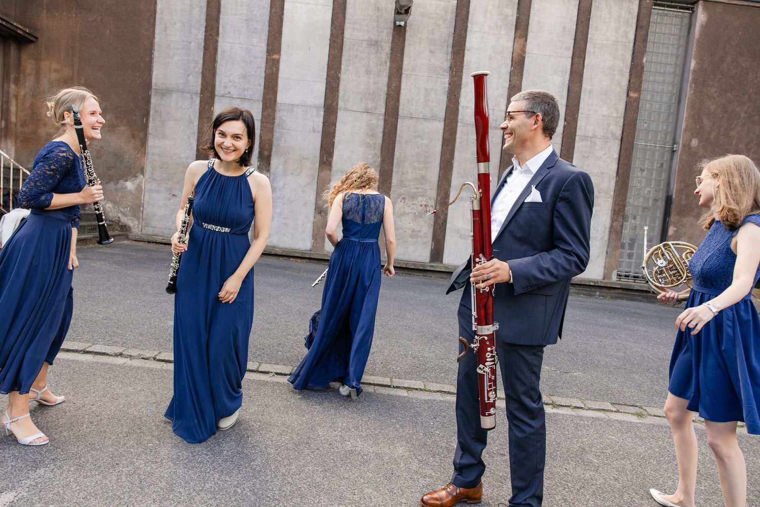 Five musicians in blue formal attire holding classical instruments outdoors, including bassoon and French horn.