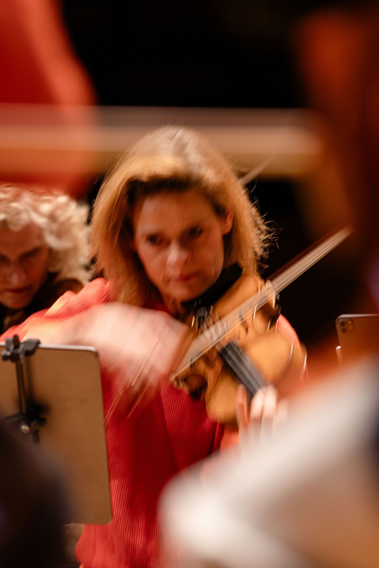 A focused female violinist in a red top performs with an orchestra, sheet music visible on a stand beside her.