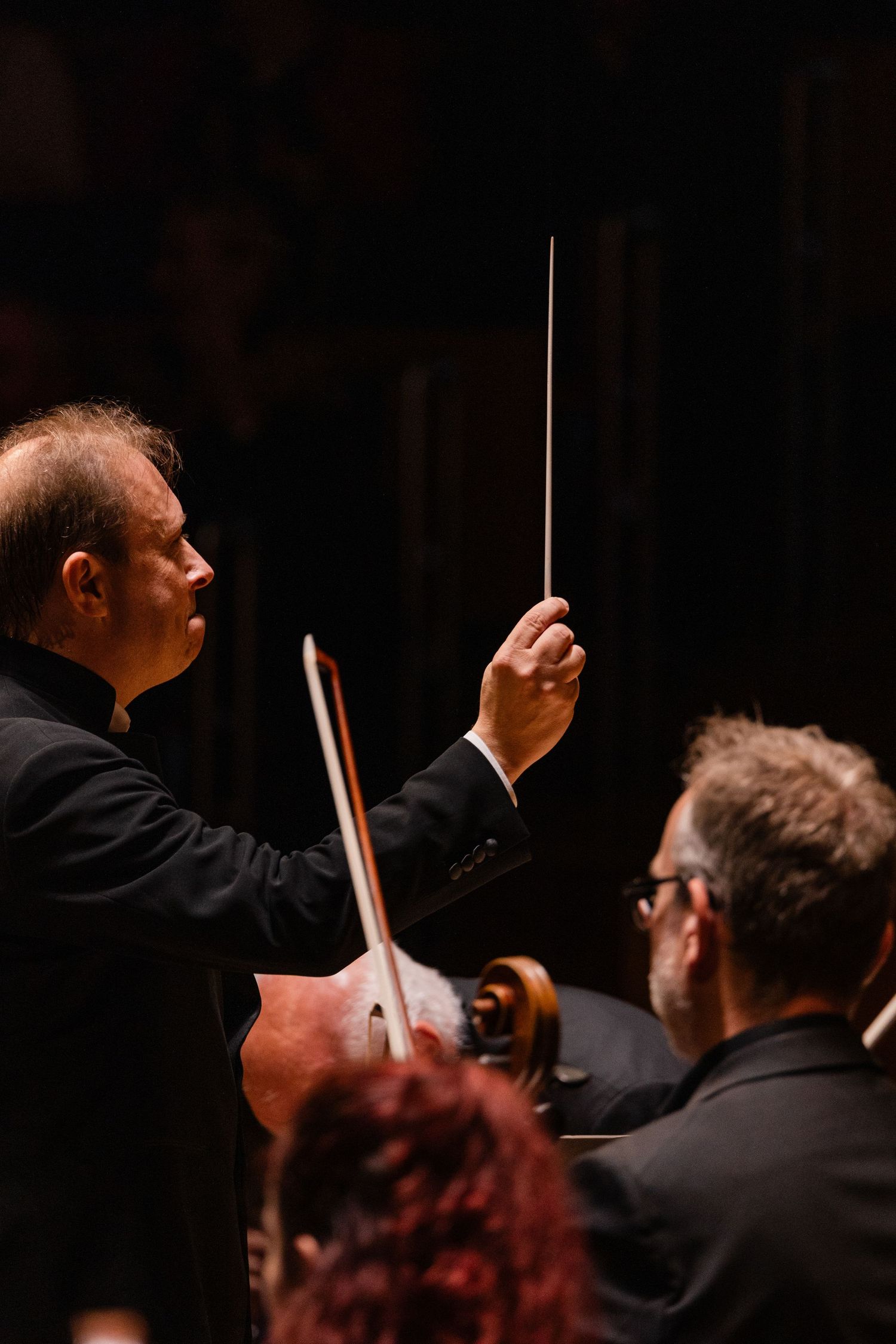 Orchestra conductor holds baton during performance in dark concert hall with dramatic lighting.