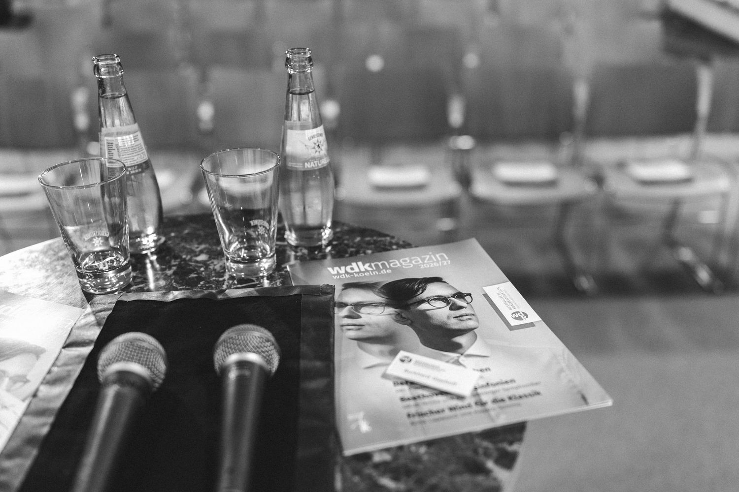 Black and white photo of microphones, water bottles, glasses, and a magazine on a conference table.