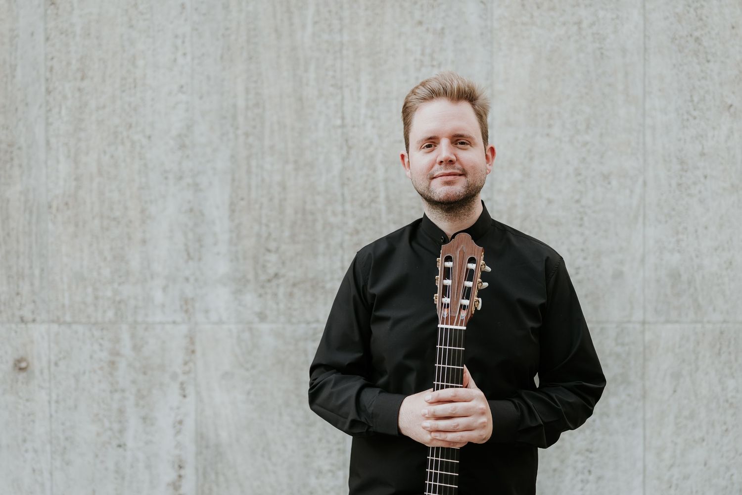 Young male guitarist in black shirt holding classical guitar against concrete wall.
