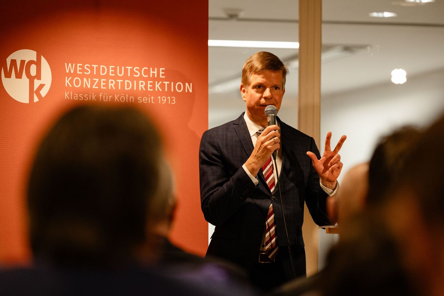 A man speaks into a microphone at a Westdeutsche Konzertdirektion event, gesturing expressively before an audience.