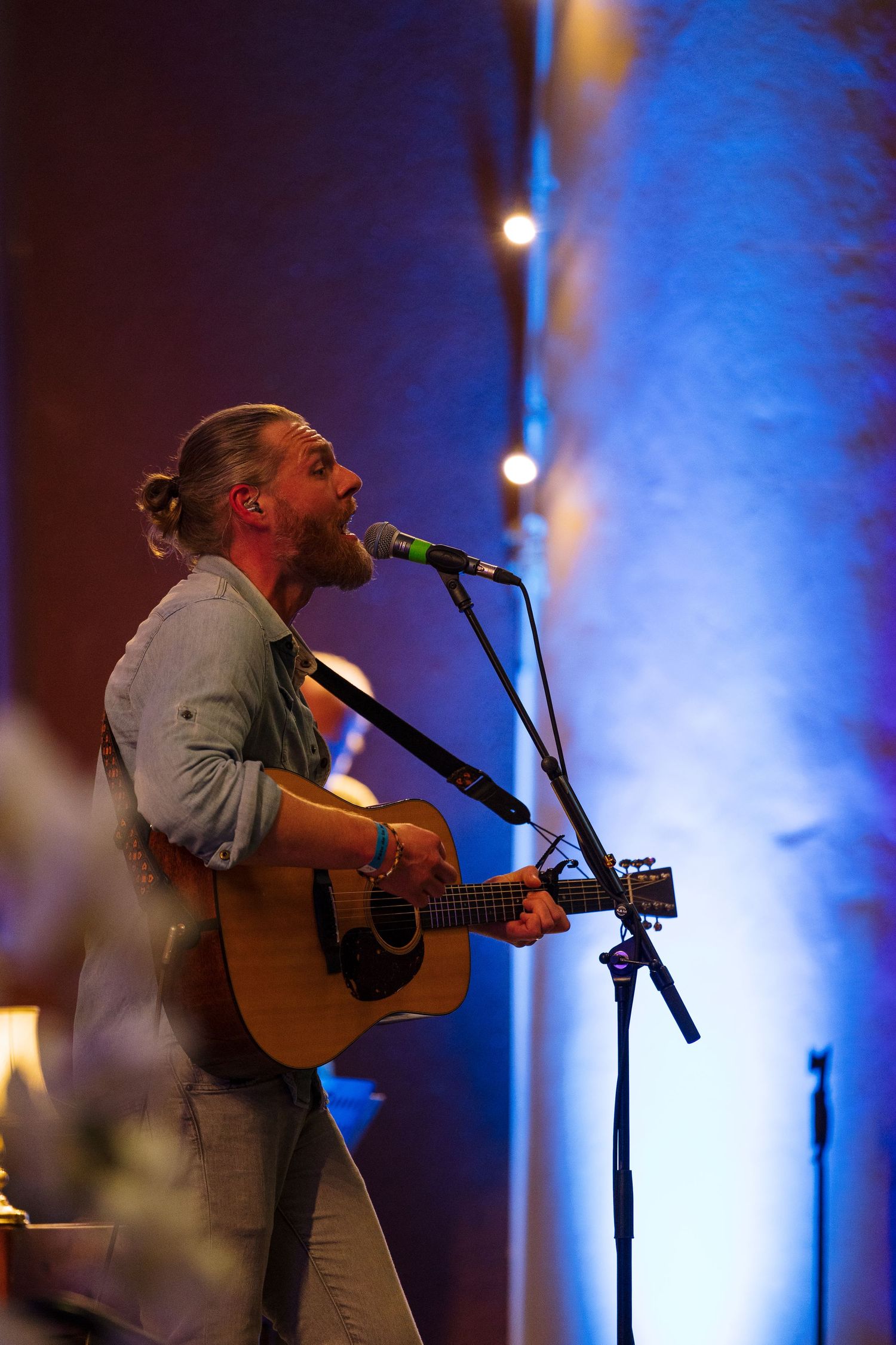 Bearded male musician with bun hairstyle playing acoustic guitar and singing into microphone on stage with blue lighting.