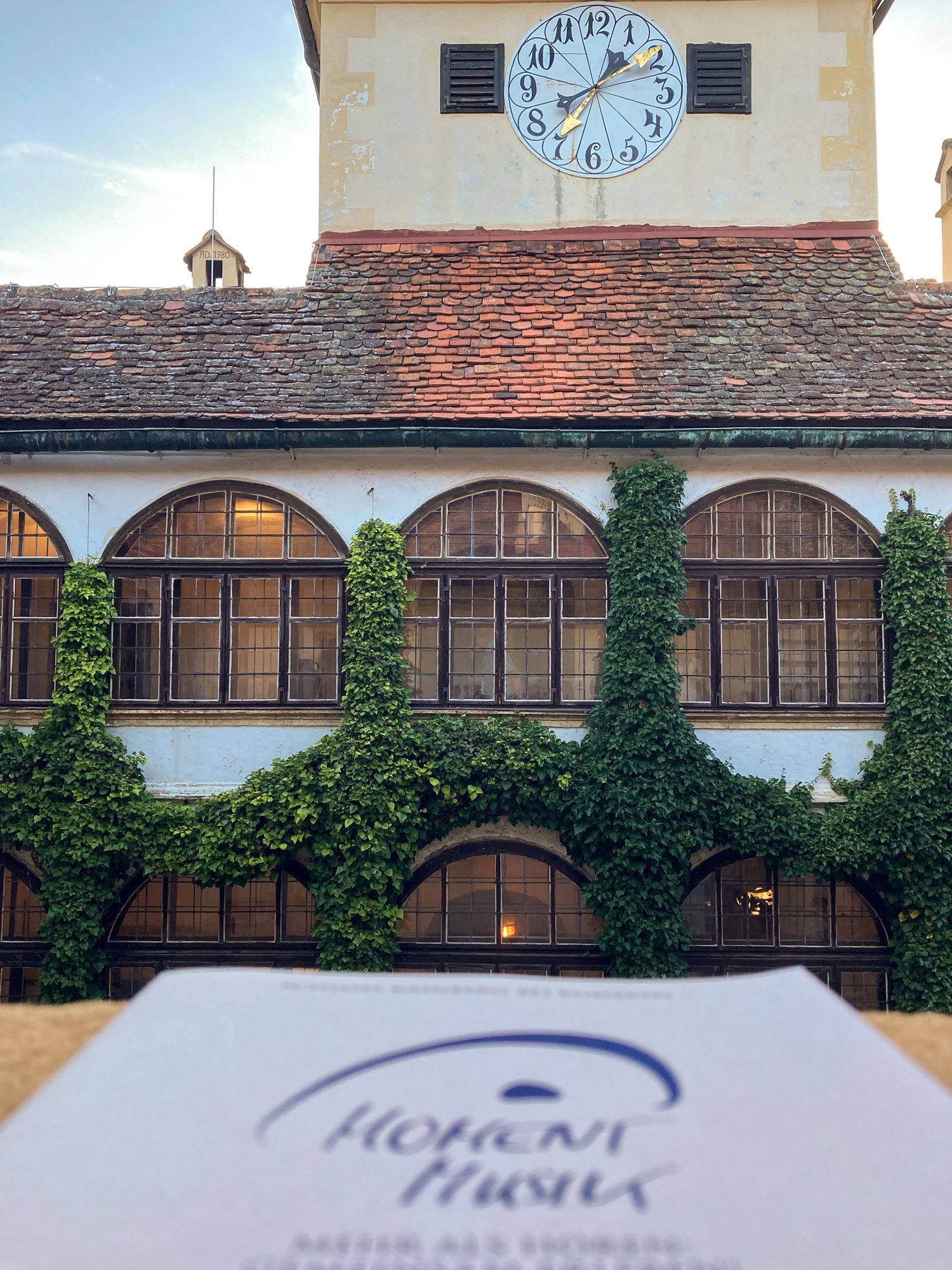 Historic building with clock tower, ivy-covered arched windows, and a Hohens Haus branded card in foreground.
